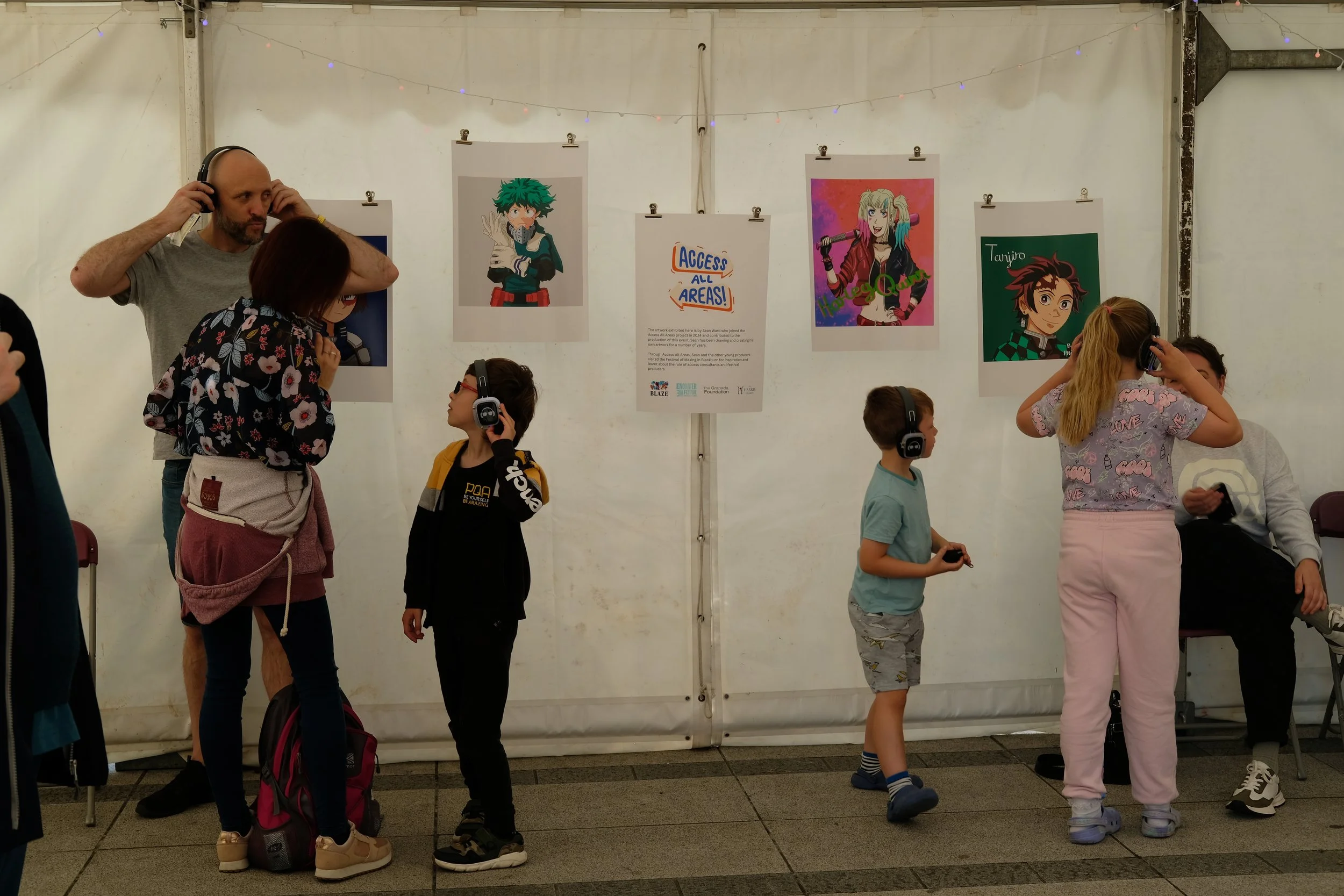 A group of children and adults at an indoor art exhibit, looking at colorful character illustrations on hanging posters, with some children wearing headphones.