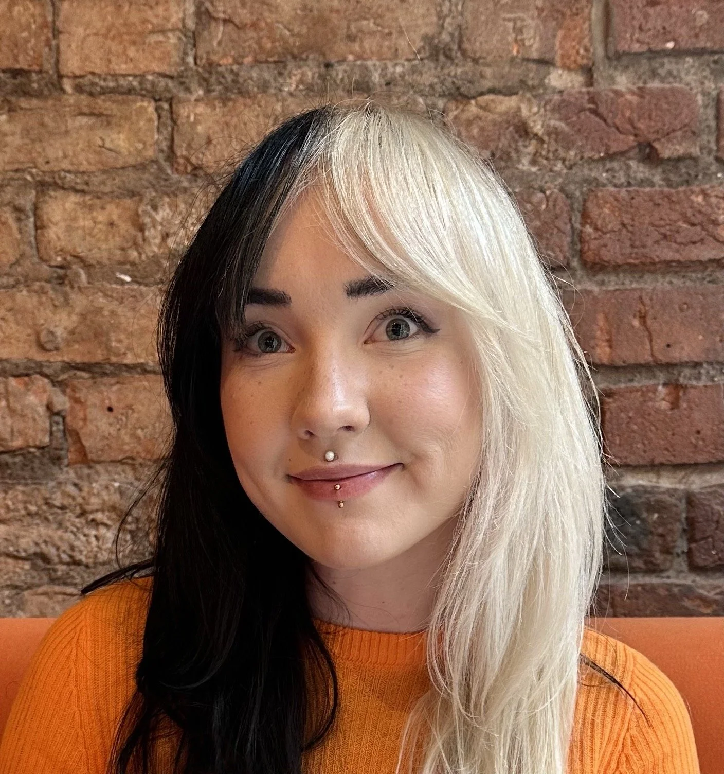 A woman with black and platinum blonde hair, wearing an orange top, smiling, with a brick wall in the background.