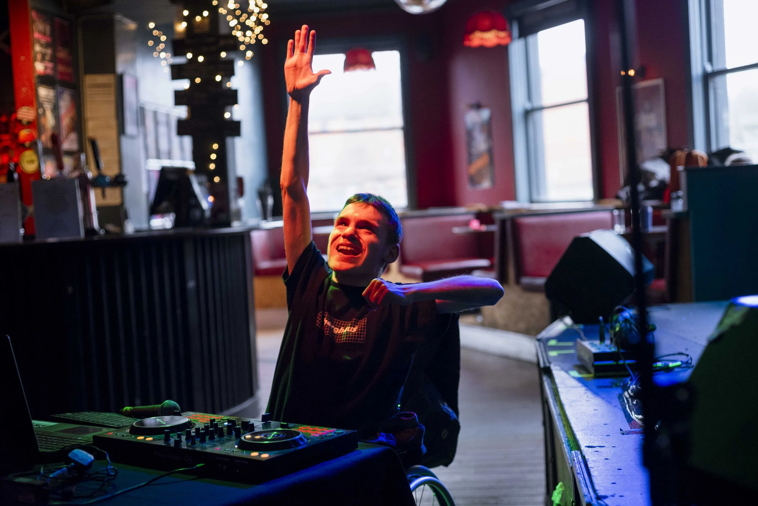 A young person with short hair smiling and raising their hand at a DJ set-up in a dimly lit room with large windows, red walls, and string lights.