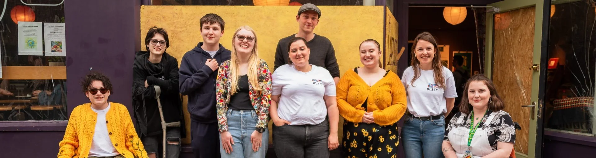 A group of nine diverse young adults standing outdoors, smiling and posing for a photo in front of a yellow wall and a purple building with glass doors, some wearing casual clothing and accessories.