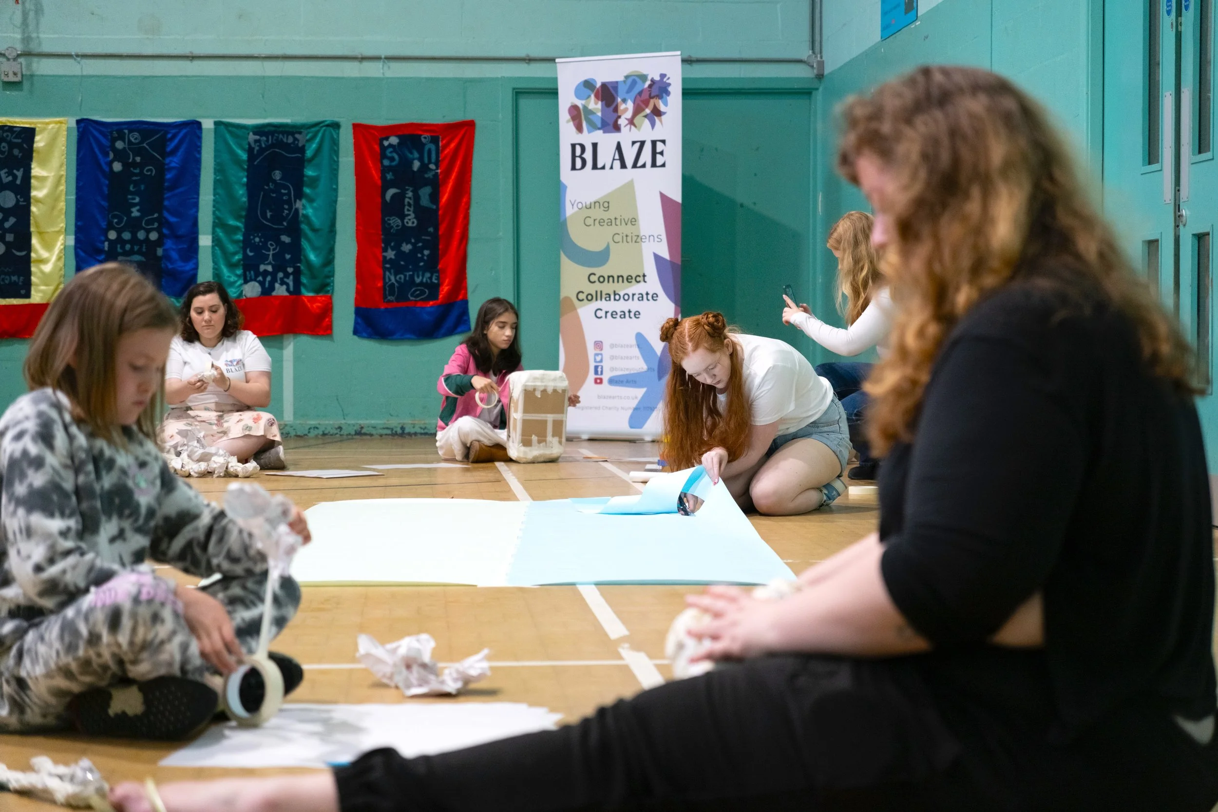 Group of children and teenagers sitting on the floor of a gymnasium, engaged in an arts and crafts activity. A colorful banner with the words 'BLAZE' and phrases like 'Young Creative Citizens,' 'Connect,' 'Collaborate,' and 'Create' stands behind them. Decorative banners hang on the wall.