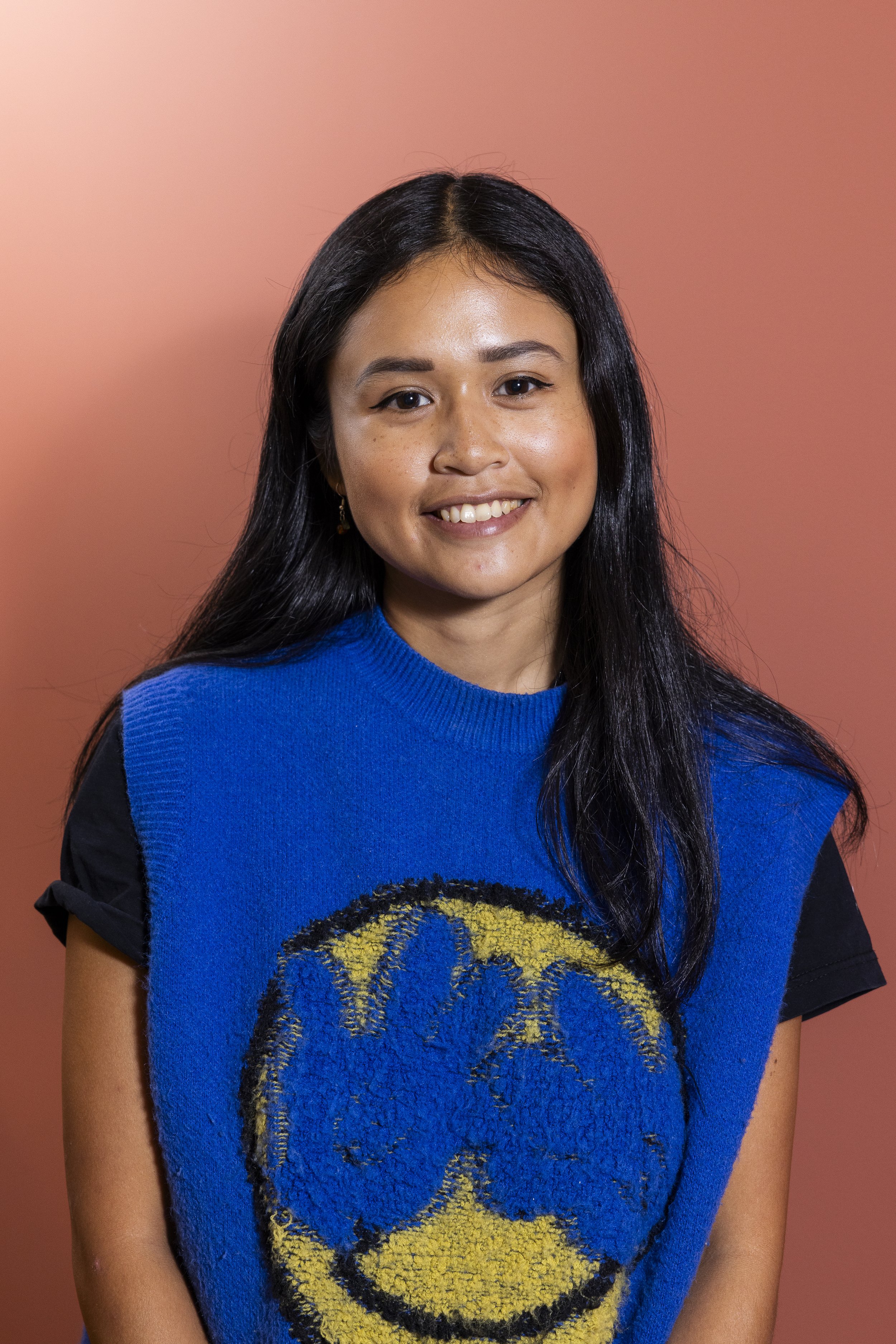 A woman with long black hair, wearing a blue sweater with a yellow smiley face on it, smiling against a pink background.