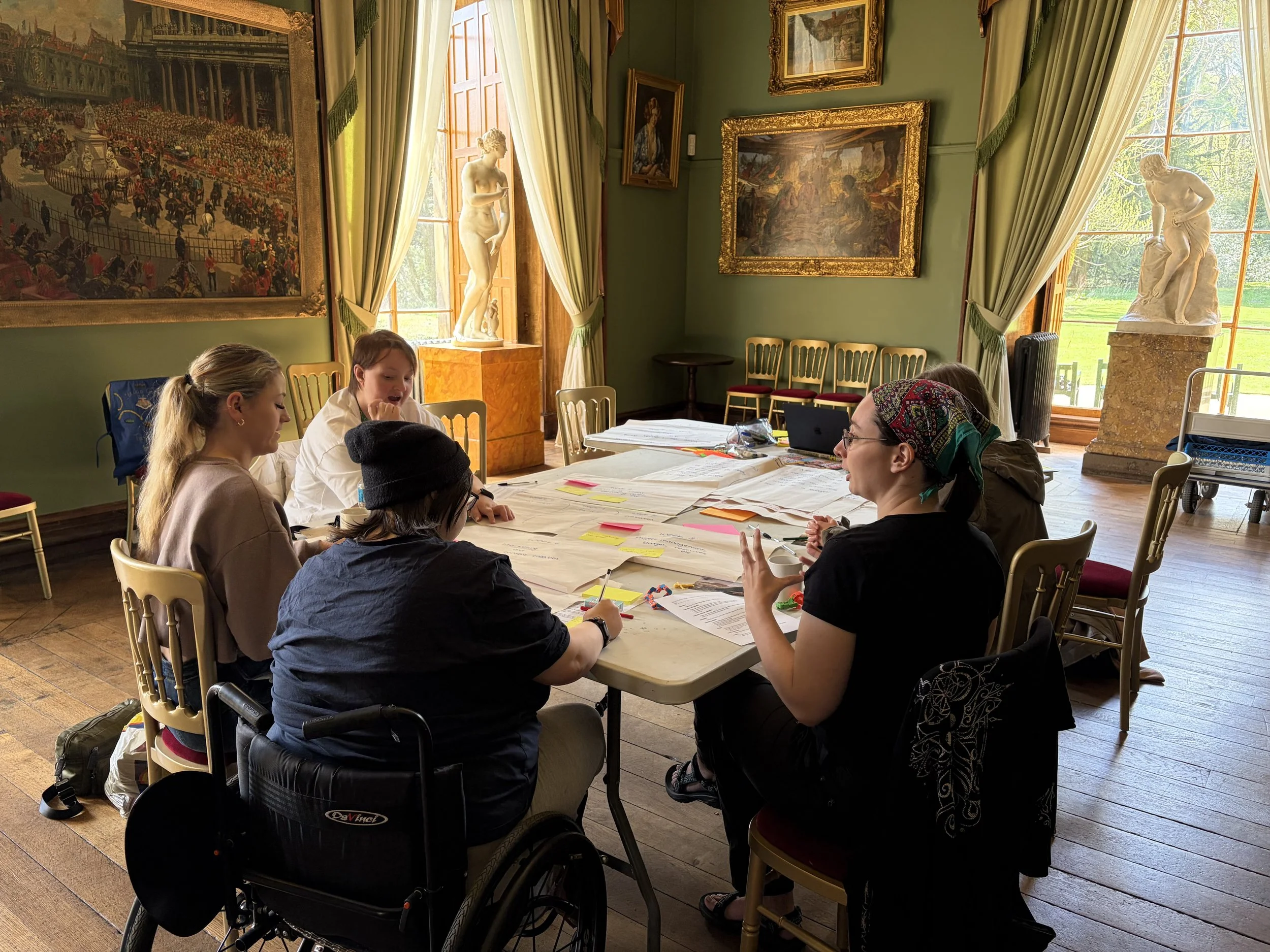 A group of people gathered around a large table in an art gallery or museum. The room has green walls with framed paintings and sculptures on pedestals. Large windows with curtains allow natural light to illuminate the space.