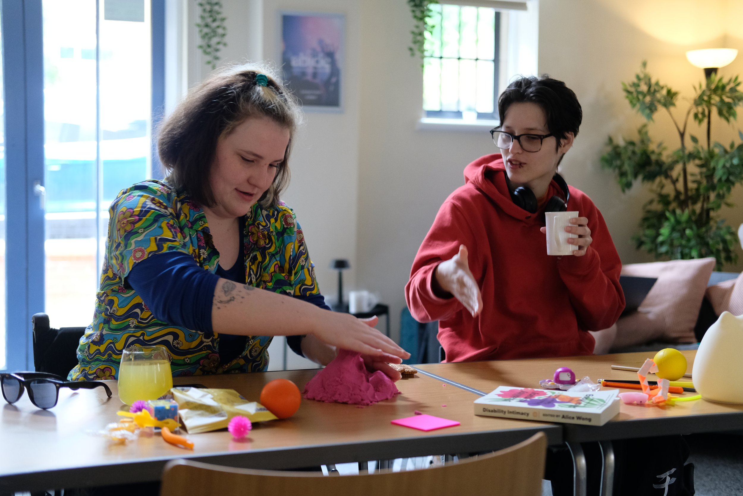 Two young women sit at a table in a bright, homey room with large windows and curtains. One woman, with short hair and glasses, wears a red hoodie and holds a white mug, while the other, with shoulder-length hair and a colorful shirt, smiles and interacts with various arts and craft supplies on the table, including eggs, paper, and toys.