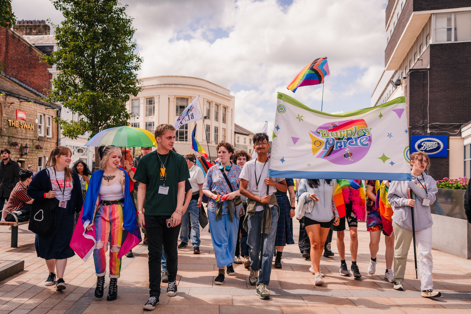 A diverse group of people walking in a pride parade holding a large banner that reads 'Burnley Pride'. Some people are holding rainbow flags and umbrellas, and wearing colorful clothing, celebrating outdoors on a city street with buildings and a cloudy sky in the background.