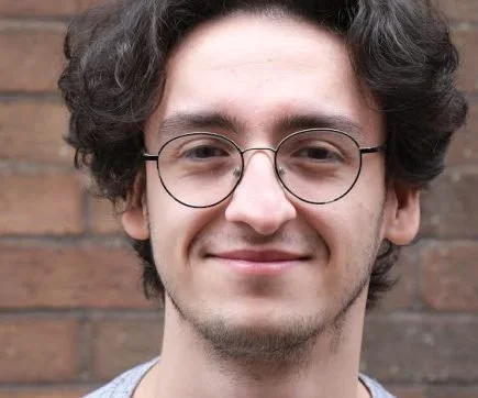 Close-up of a young man with curly hair, wearing glasses, smiling, standing in front of a brick wall.