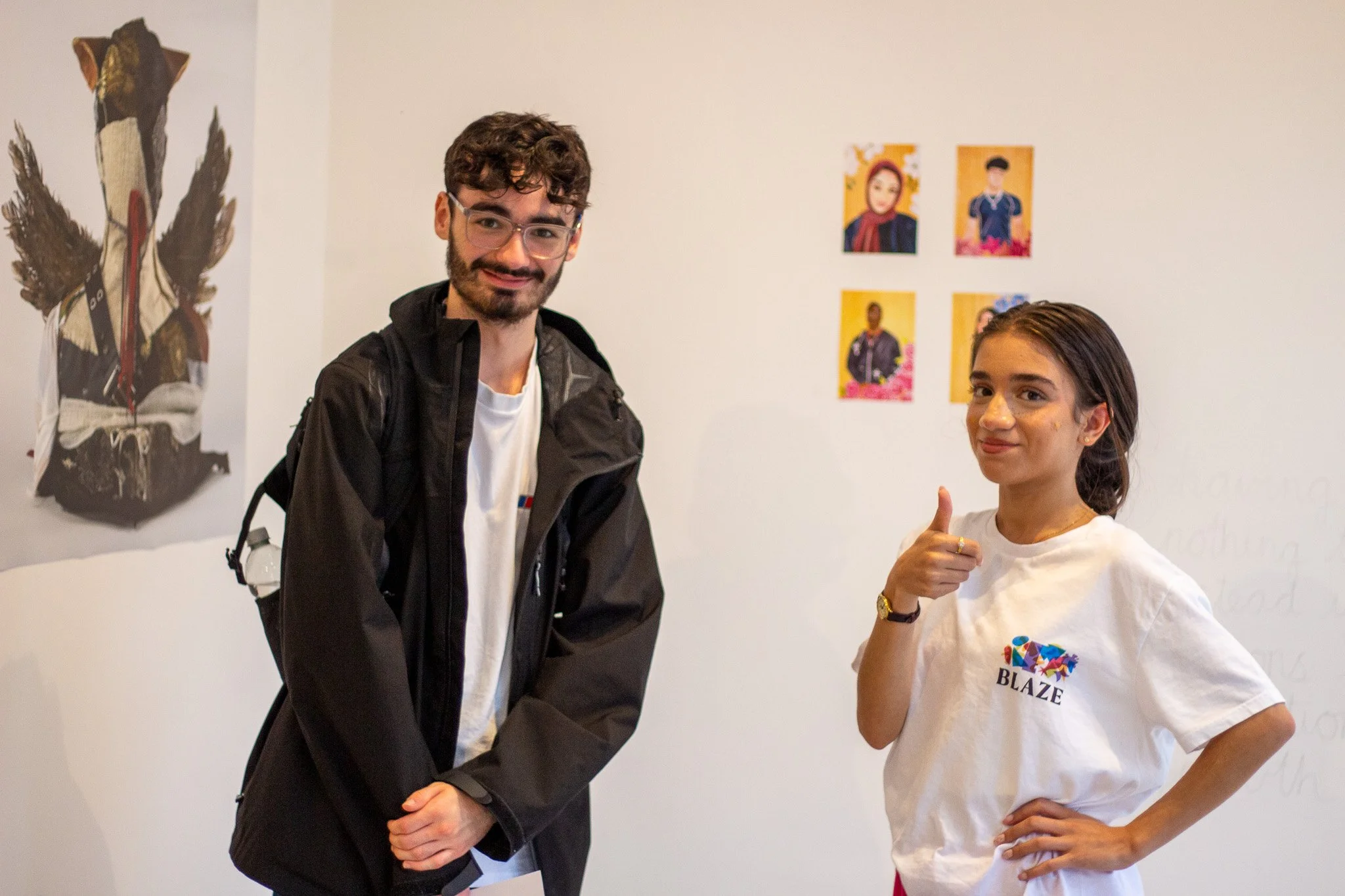 A man with glasses and a woman with long brown hair pose together in front of a white wall decorated with colorful artwork. The man is wearing a black jacket and white shirt, smiling at the camera. The woman is wearing a white T-shirt with a colorful graphic and the text 'BLAZE', smiling and giving a thumbs up.