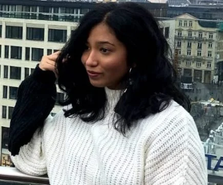 Woman with dark curly hair wearing a white sweater, standing near a window with city buildings in the background.