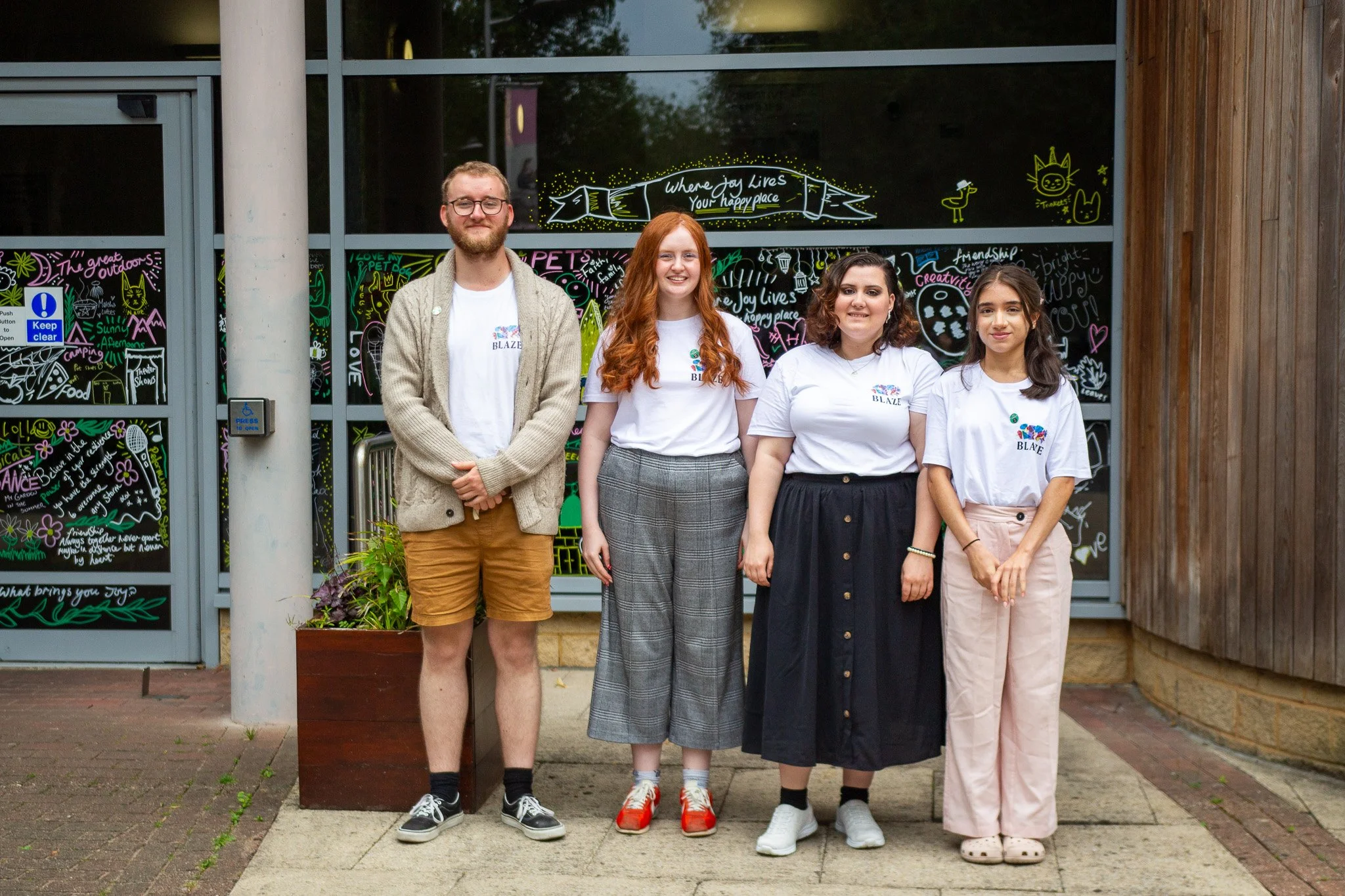 Four young adults standing outdoors in front of a glass window decorated with colorful drawings and writings. The group includes three women and one man, all smiling at the camera. The man is wearing glasses, a beige cardigan, a white t-shirt, and tan shorts. The woman next to him has long red hair, a white t-shirt, and gray checkered pants. The third woman has curly dark hair and is wearing a white t-shirt and a black skirt with wooden buttons. The fourth woman has long dark hair, a white t-shirt, and light pants. Behind them, handwritten messages include phrases like 'Where joy lives,' 'Your happy place,' and various doodles.