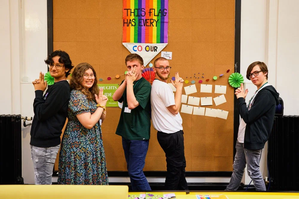Five young people standing in front of a bulletin board with rainbow decorations and a sign that says 'This flag has every color.' They are posing with playful expressions, some with hands in funny positions, and appear to be celebrating diversity.