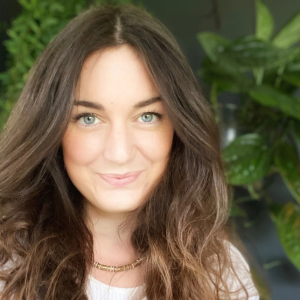A woman with long brown hair and blue eyes smiling at the camera, with green plants in the background.
