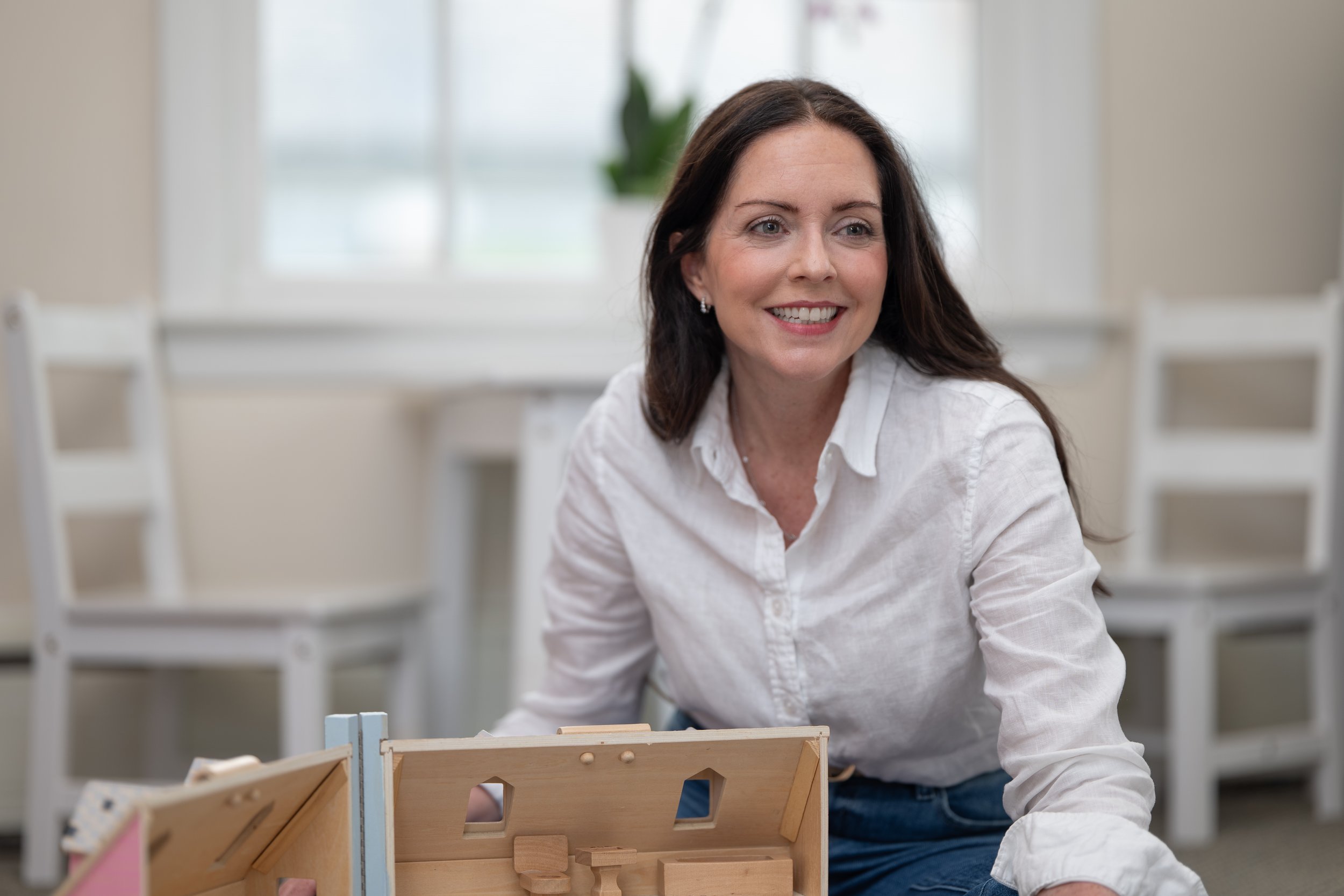 Smiling woman with dark brown hair in a white shirt sitting at a table with wooden models.