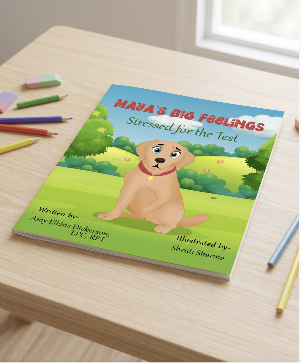 Children's book titled "Maya's Big Feelings: Stressed for the Test" on a light wooden table with colored pencils and erasers nearby, featuring a cartoon puppy on the cover in a green outdoor setting.