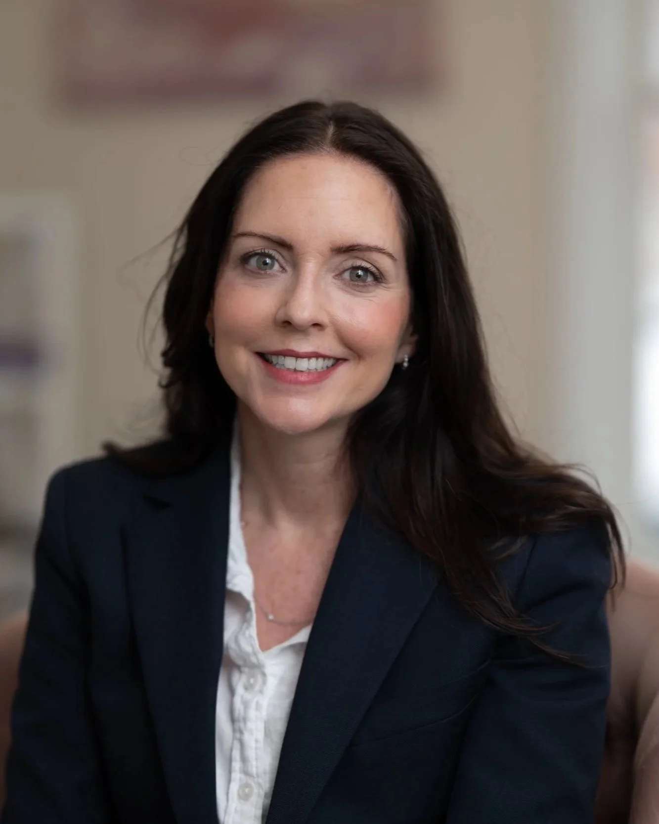 A woman with long dark hair, blue eyes, and a bright smile, wearing a dark suit and white blouse, posing for a professional portrait indoors.