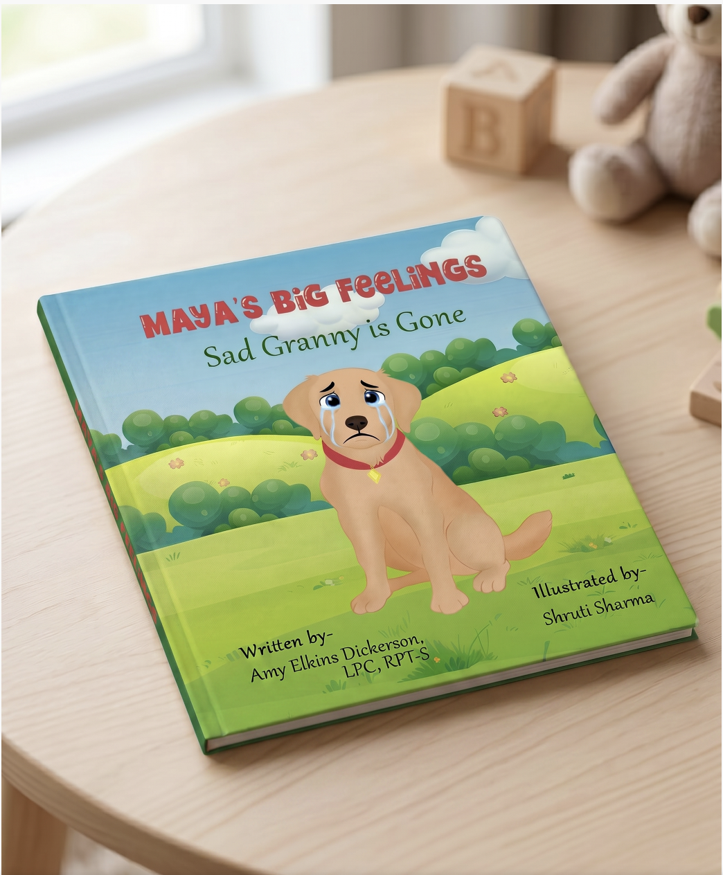 Children's children's book titled 'Maya's Big Feelings: Sad Granny is Gone' featuring a cartoon sad puppy with teary eyes sitting on grass with lush green bushes and a clear blue sky with white clouds in the background. The book is on a wooden table with a teddy bear and wooden toy blocks nearby.