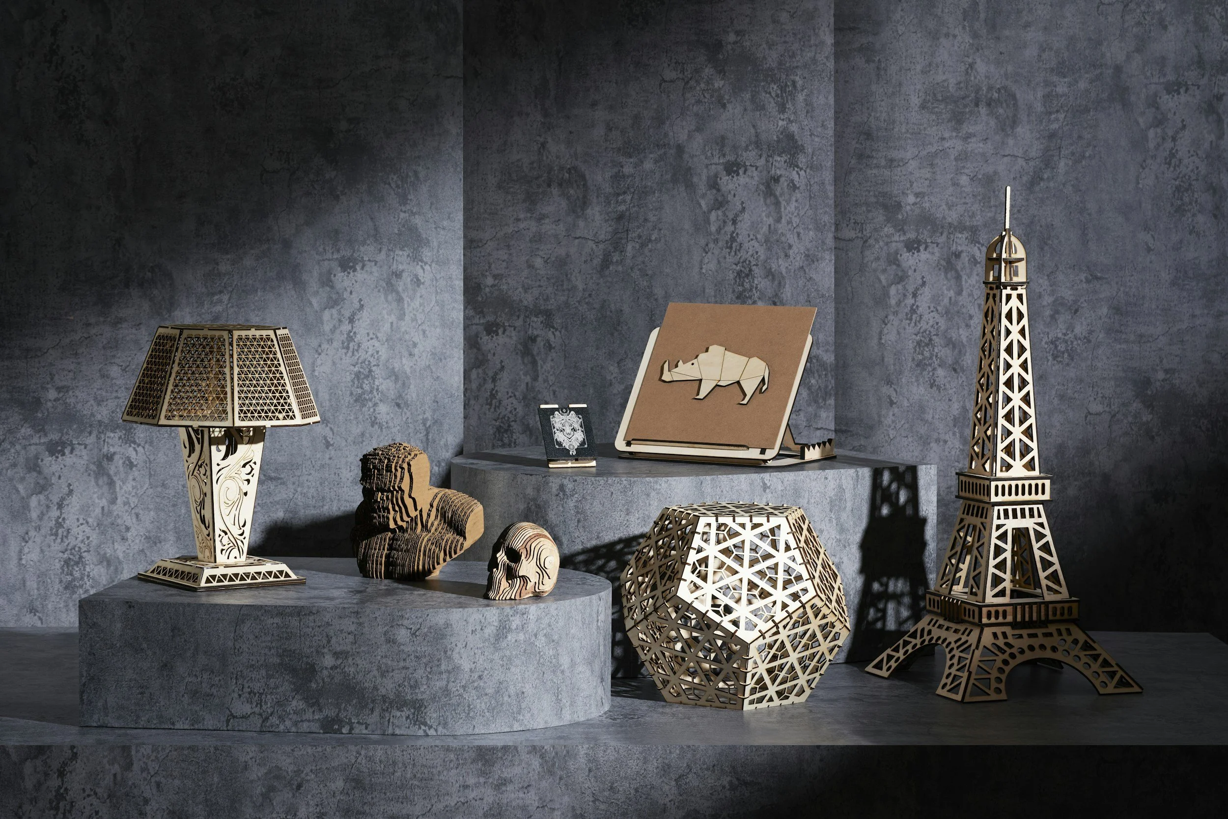 Decorative objects including a wooden table lamp, wooden skull sculptures, a notebook with a wooden rhino, a small framed photo, and various geometric wooden sculptures resembling buildings and the Eiffel Tower, all arranged on a round stone pedestal against a textured gray wall.