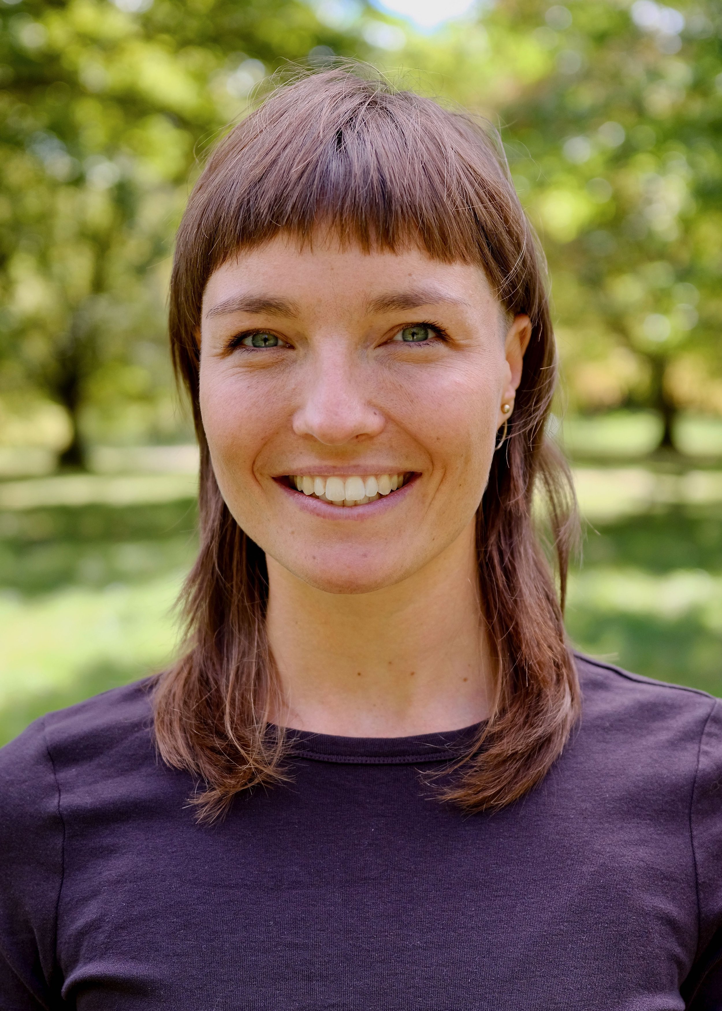 Porträt einer lachenden Frau mit braunen Haaren und grünen Augen, im Freien vor einer grünen, bewaldeten Hintergrund, trägt ein dunkelviolettes T-Shirt.