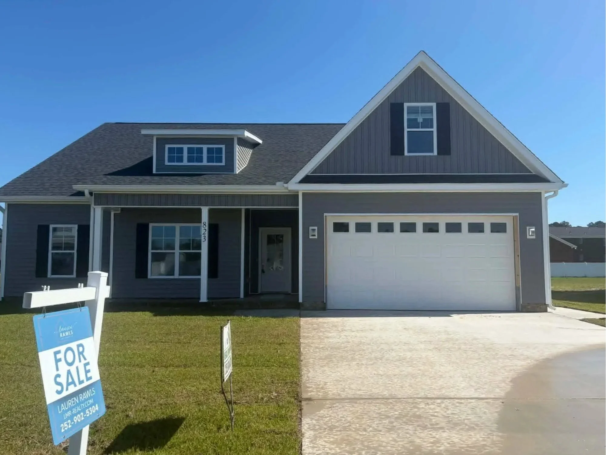 A modern house with gray siding, black shutters, and a three-car garage. A front yard has a 'For Sale' sign with contact information. The sky is blue and clear.
