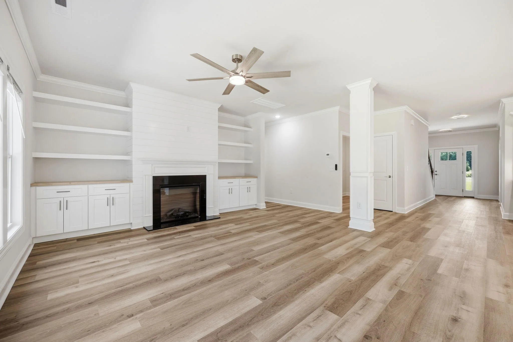 Empty living room with light-colored wood flooring, white walls, built-in white cabinets with shelves near a fireplace, ceiling fan, and front door visible in the background.