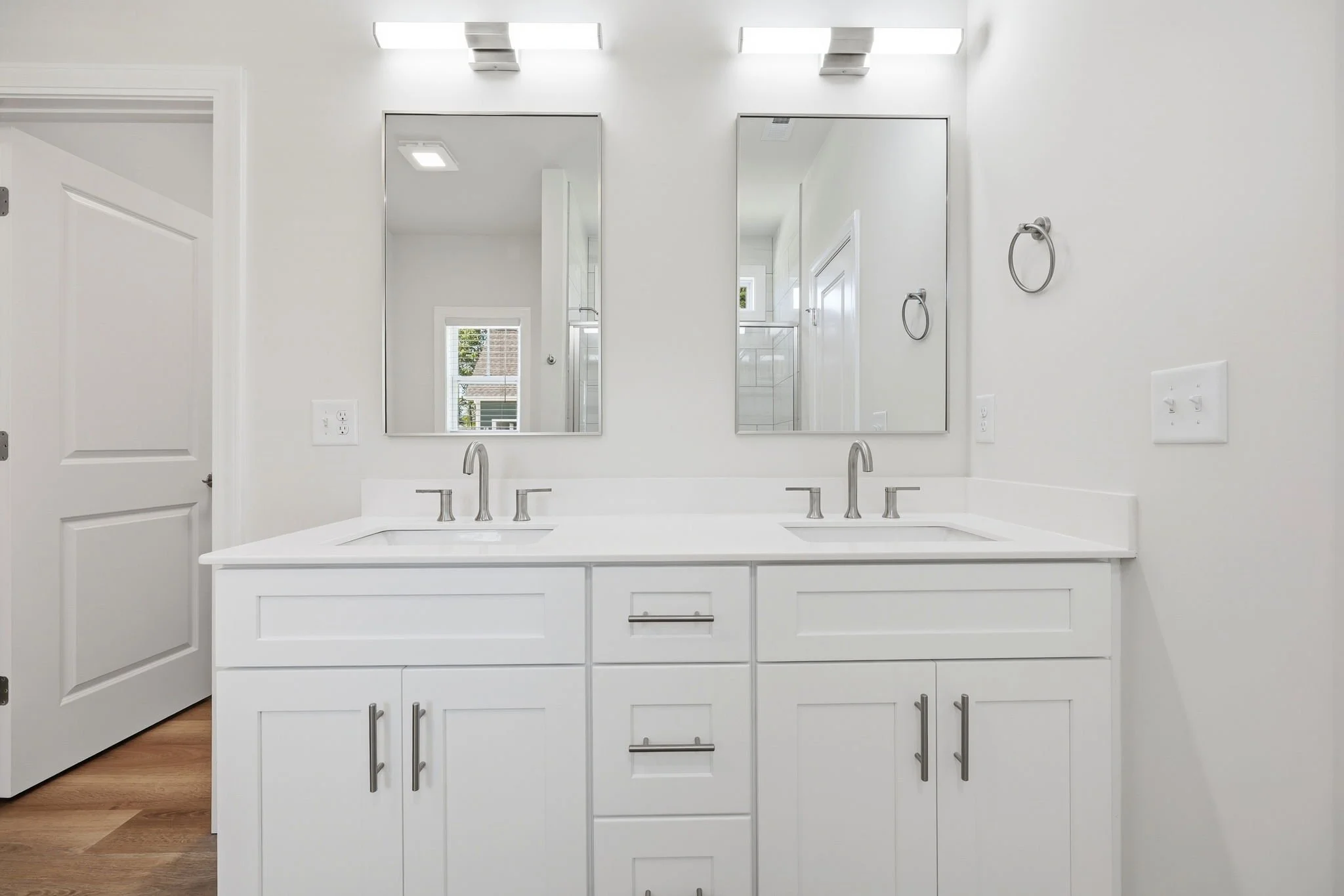 White bathroom vanity with two sinks, two mirrors, and modern silver faucets. Above mirrors are two contemporary light fixtures. The wall is painted white with a towel ring on the right and electrical outlets below.