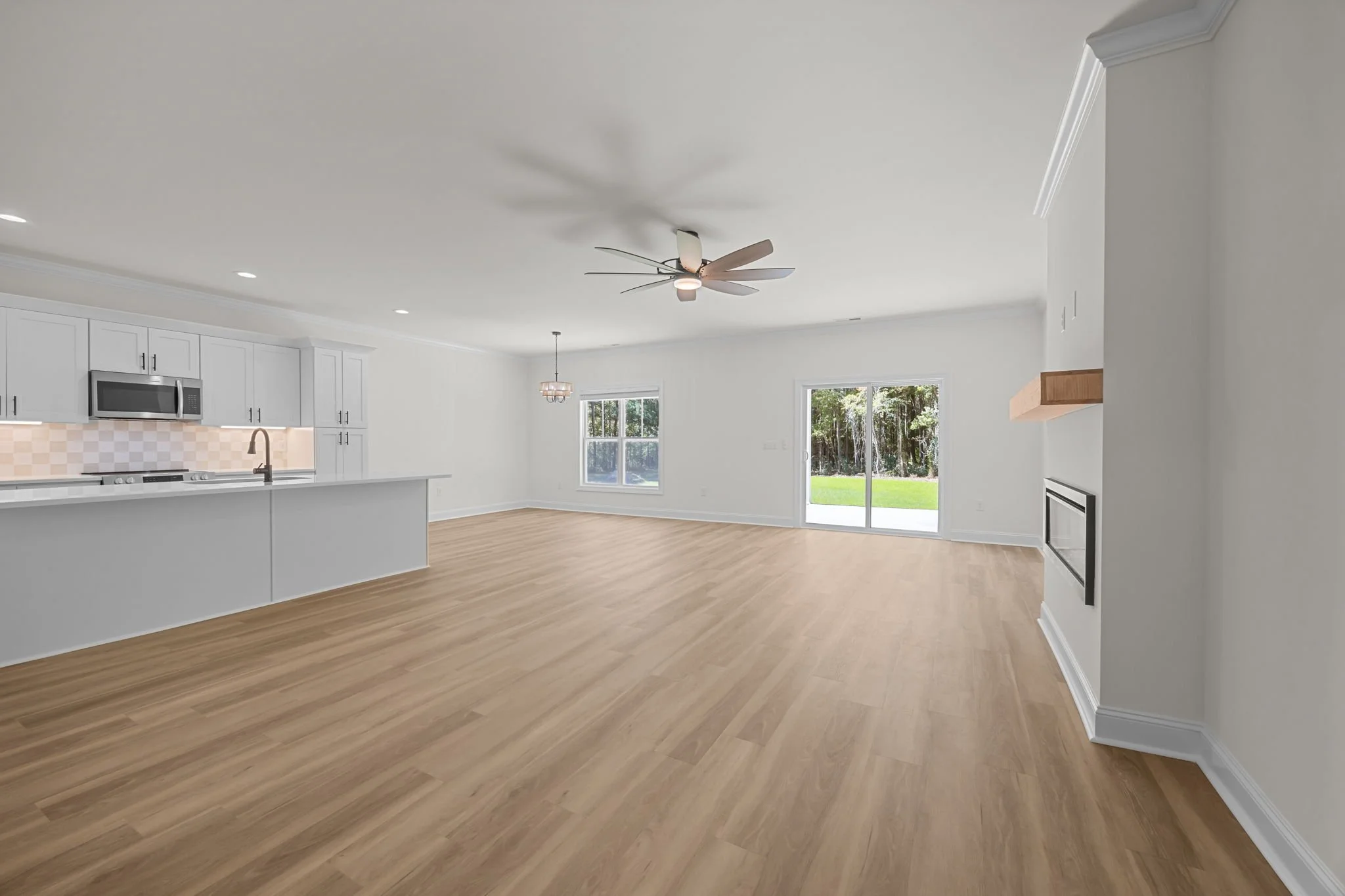 Empty open-concept living room with white walls, wooden flooring, a ceiling fan, large windows with view of green backyard, and a glimpse of kitchen with white cabinets and stainless steel appliances.