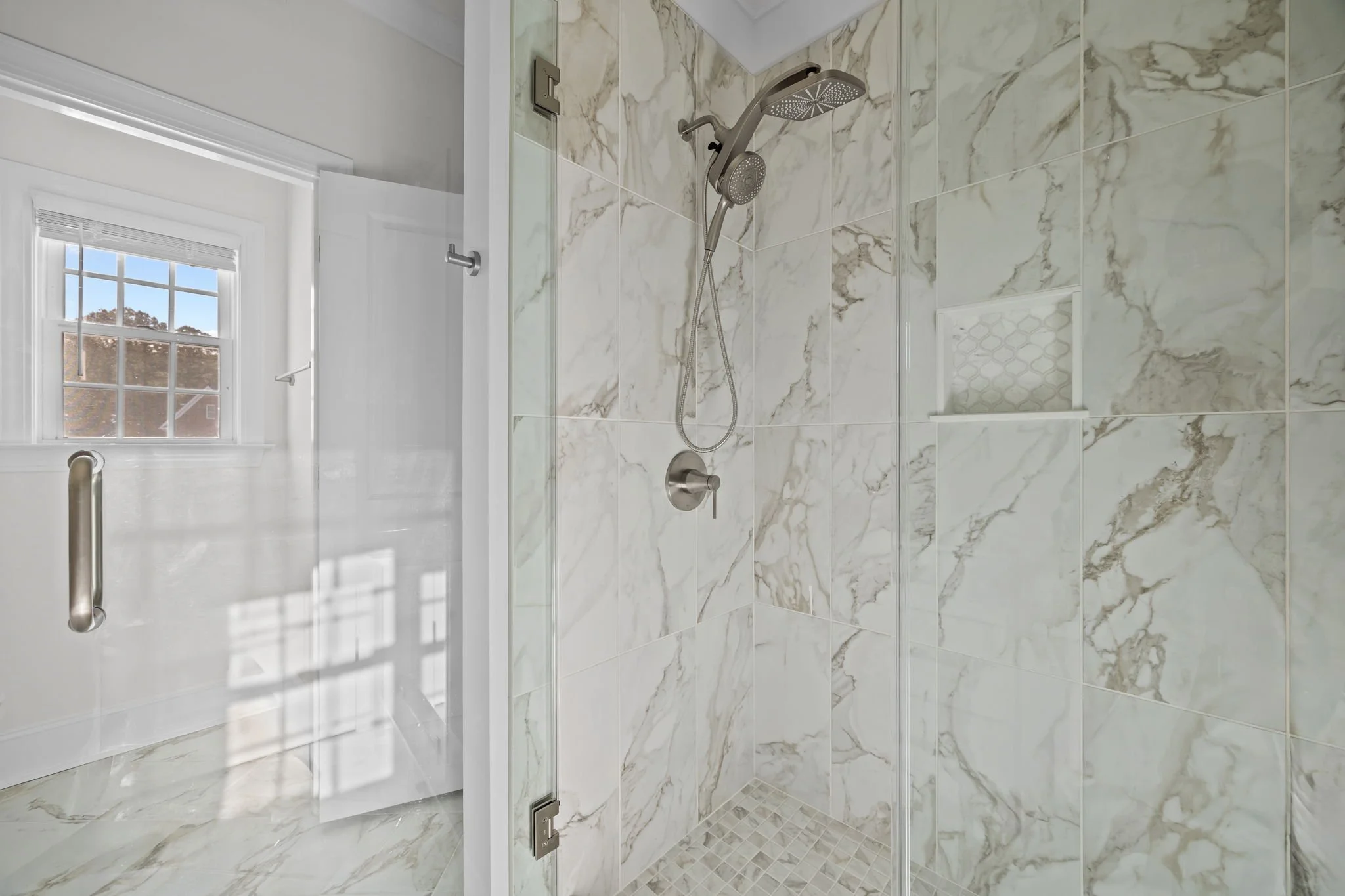 A walk-in shower with marble-like tiled walls and a glass door, featuring a rainfall showerhead and a handheld shower. 