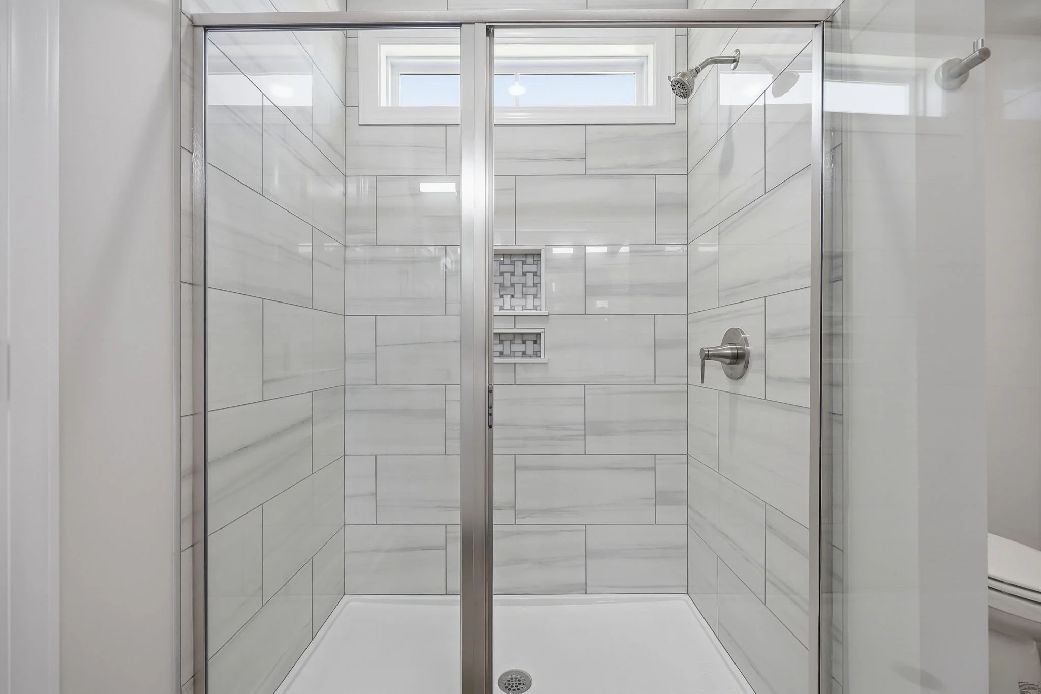 A modern shower with glass doors, marble tile walls, and built-in shelves, with a window near the ceiling for natural light.