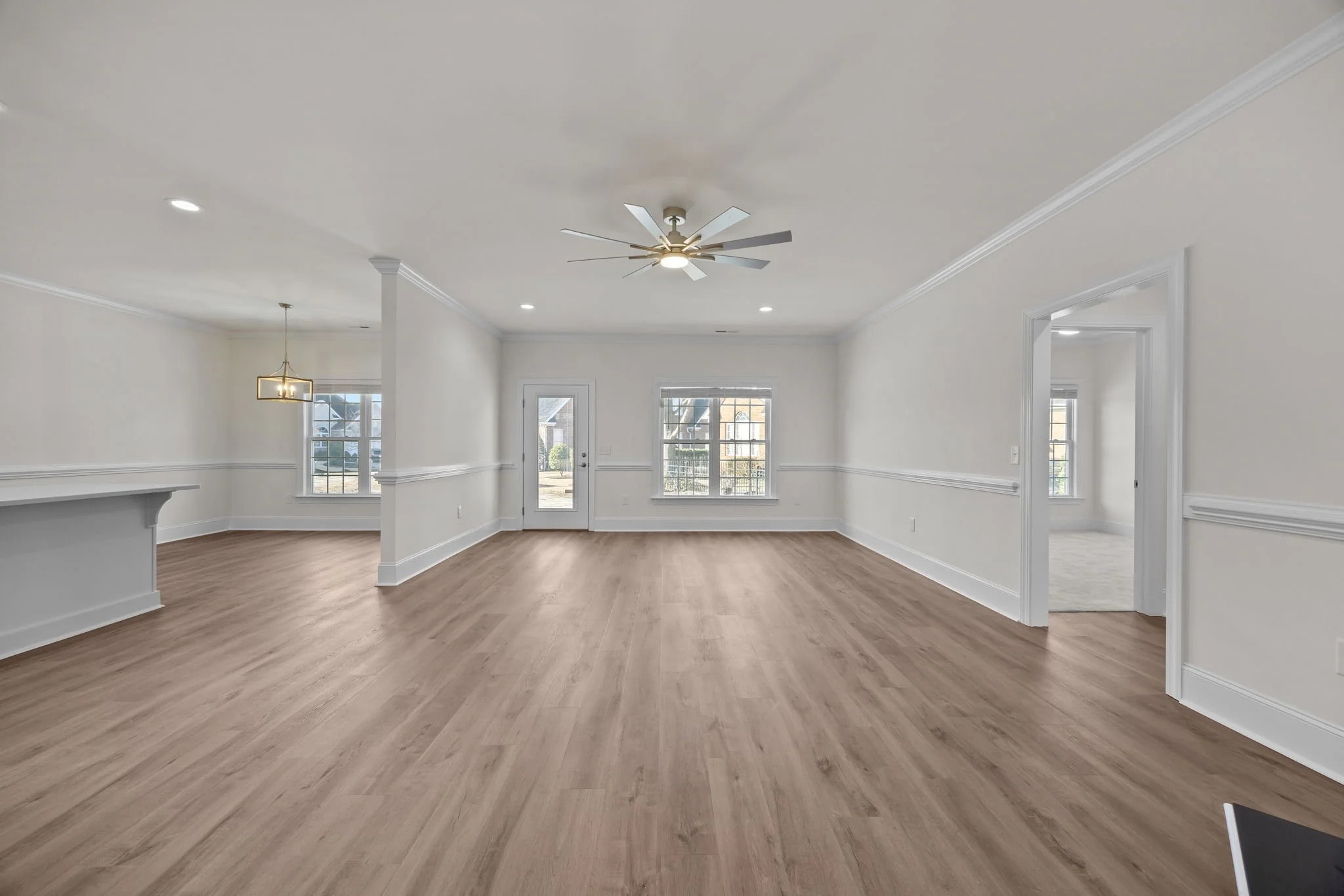 Empty living room with white walls, hardwood flooring, large windows, ceiling fan, and recessed lighting.