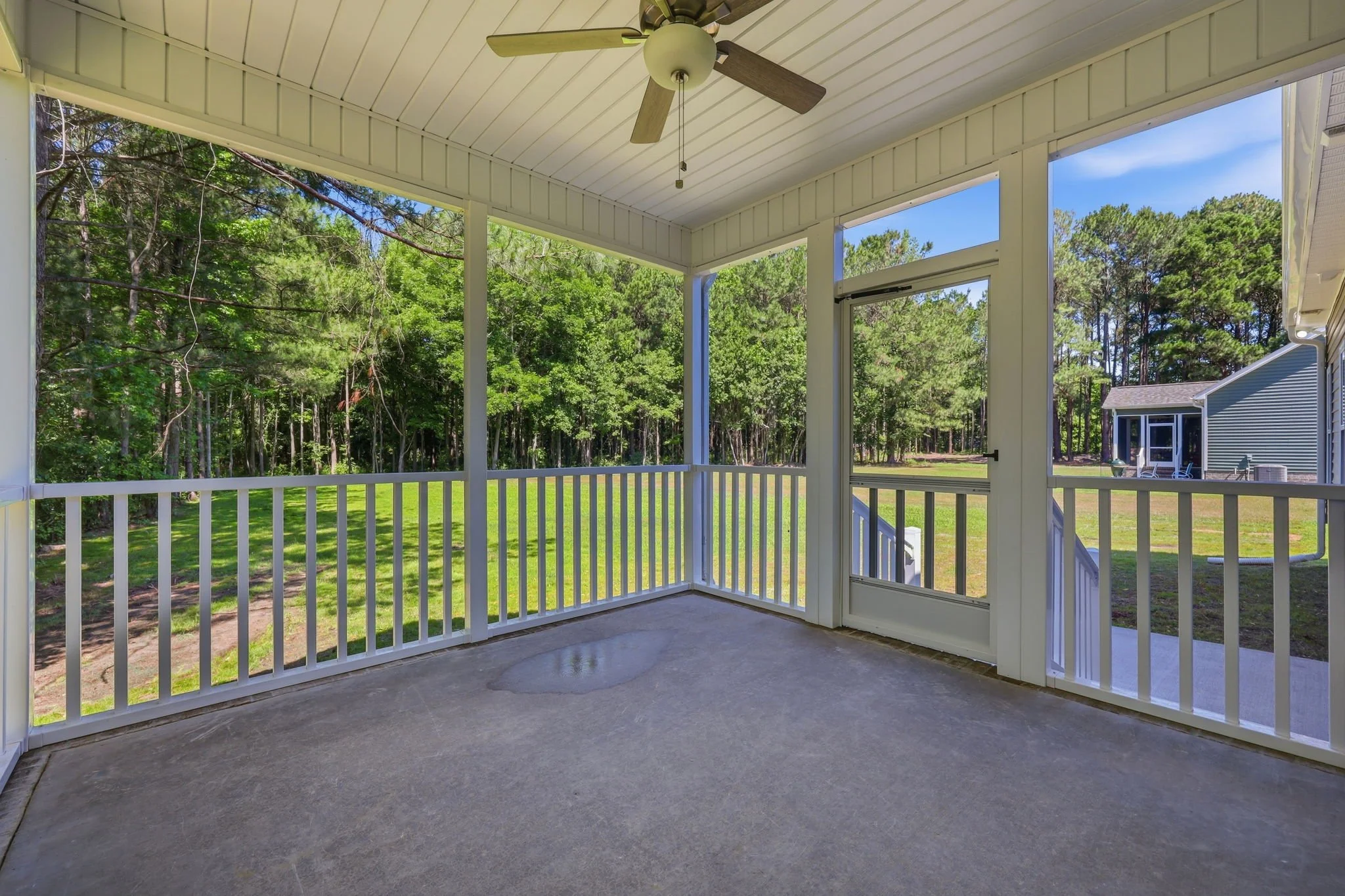 Covered porch or balcony with white railing and ceiling fan, overlooking a green backyard with trees and neighboring houses, bright blue sky.