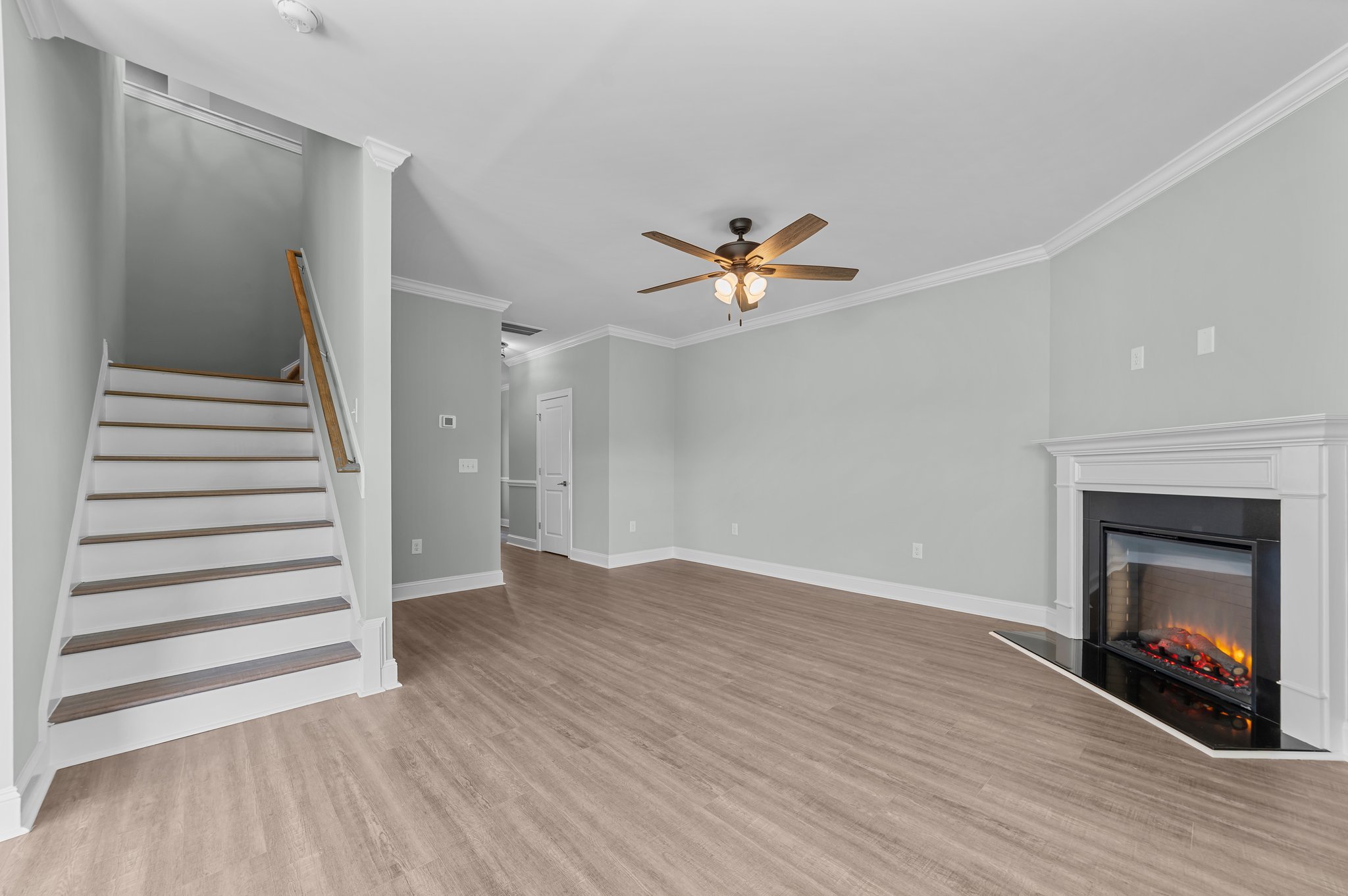 Living room with wood flooring, light gray walls, a white fireplace with a black interior, a ceiling fan with wooden blades and lights, and a staircase with white risers and wooden treads.