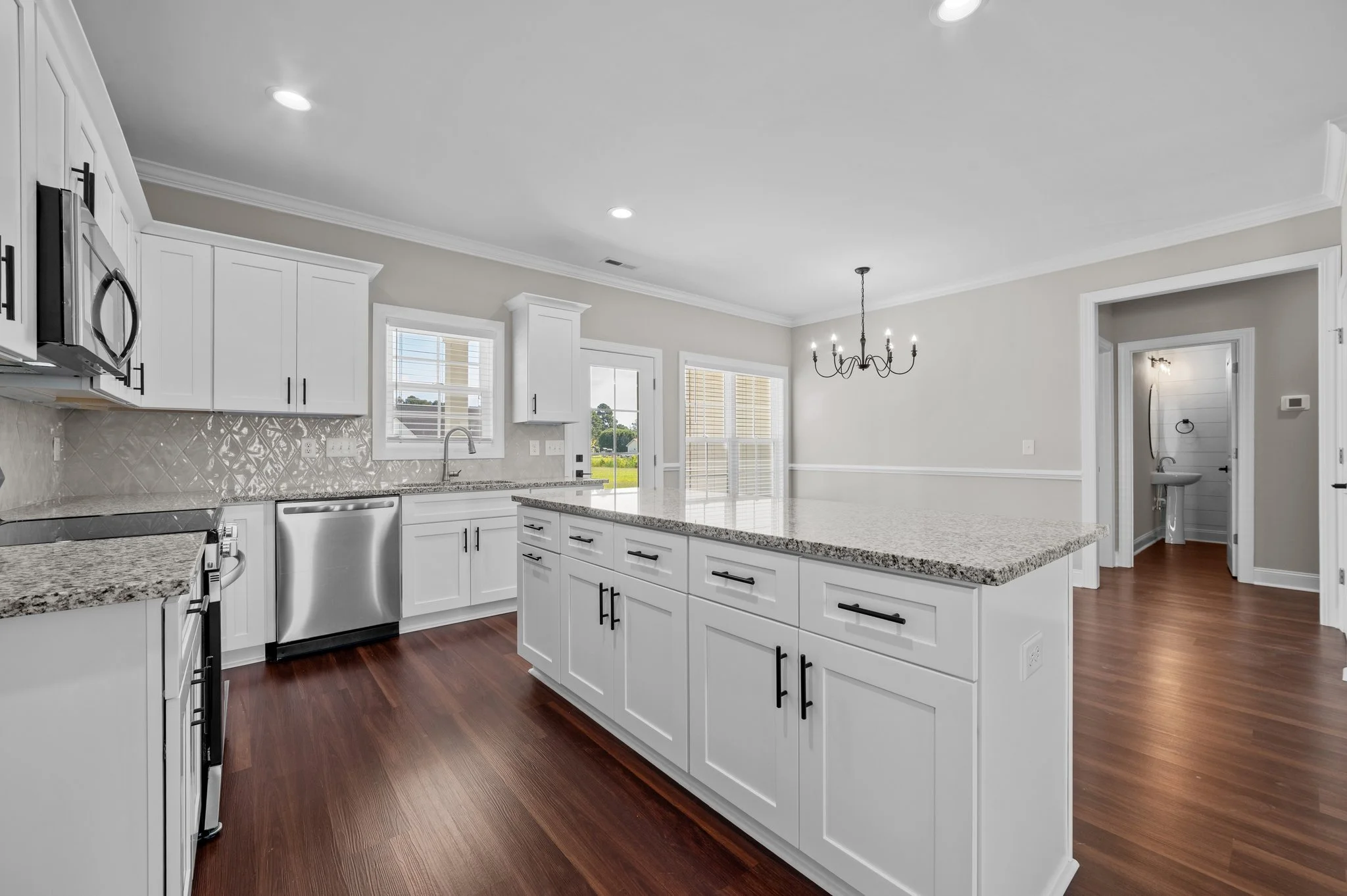 Modern kitchen with white cabinets, granite countertops, stainless steel appliances, and hardwood floors, with light neutral wall color, windows, and a dining area with a chandelier.