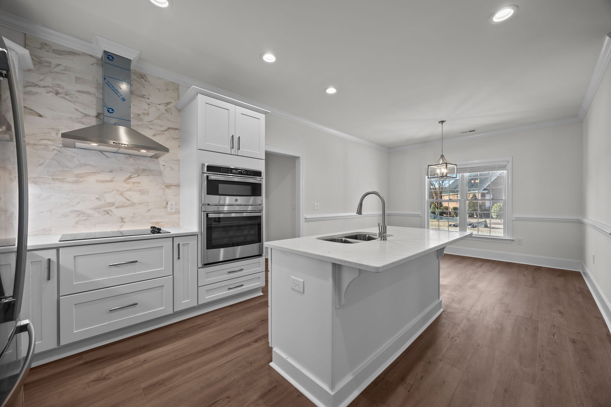 Modern kitchen with white cabinets, stainless steel appliances, a marble backsplash, a kitchen island with a sink, and a large window letting in natural light.