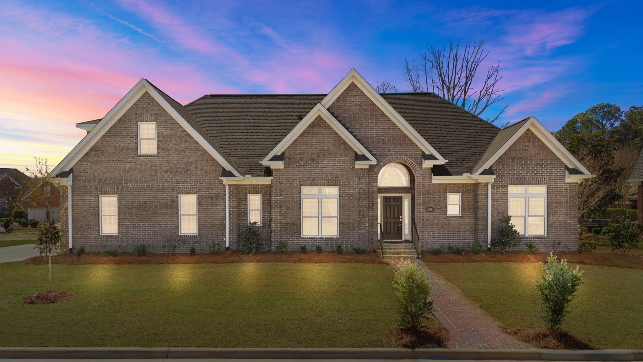 A two-story brick house with a gabled roof and front porch, situated on a well-maintained lawn with small trees and shrubs, under a colorful sky at dusk.