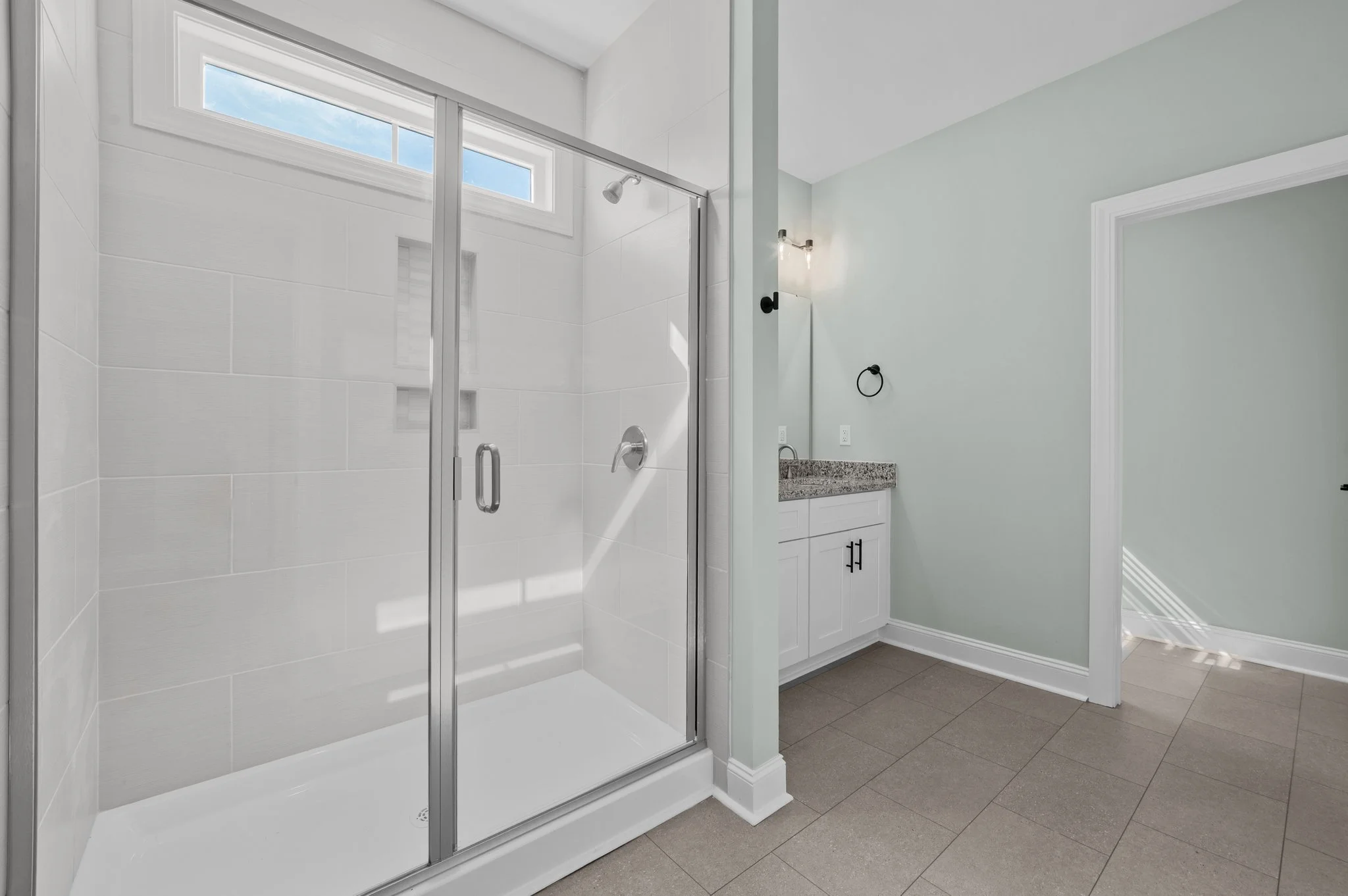 Modern bathroom with a glass-enclosed shower featuring white tiled walls, a small horizontal window, a showerhead, and a vanity with a granite countertop and cabinet beneath it.