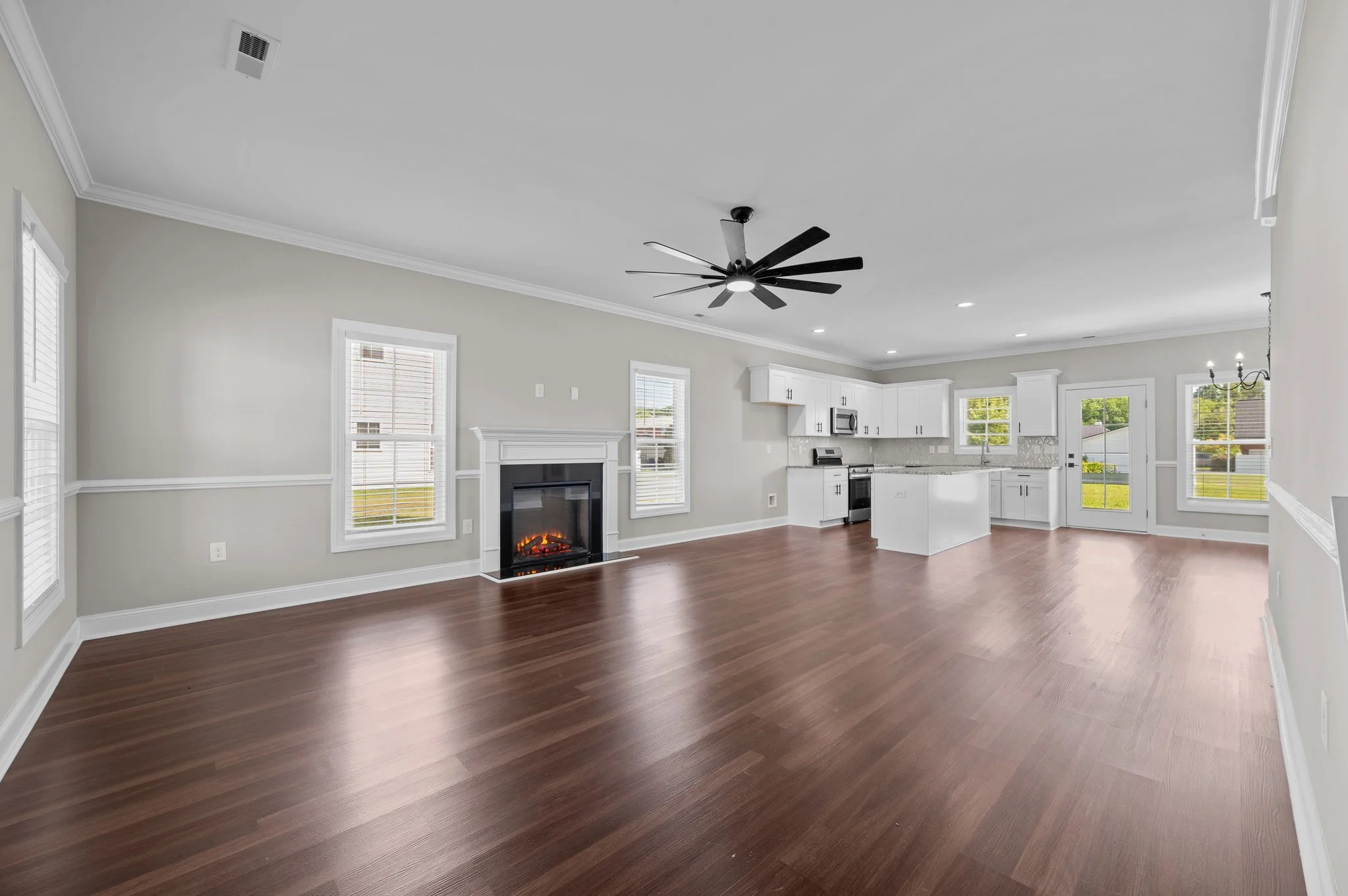 Open-concept living room and kitchen with white cabinets, dark wood flooring, ceiling fan, fireplace, and multiple windows with blinds.