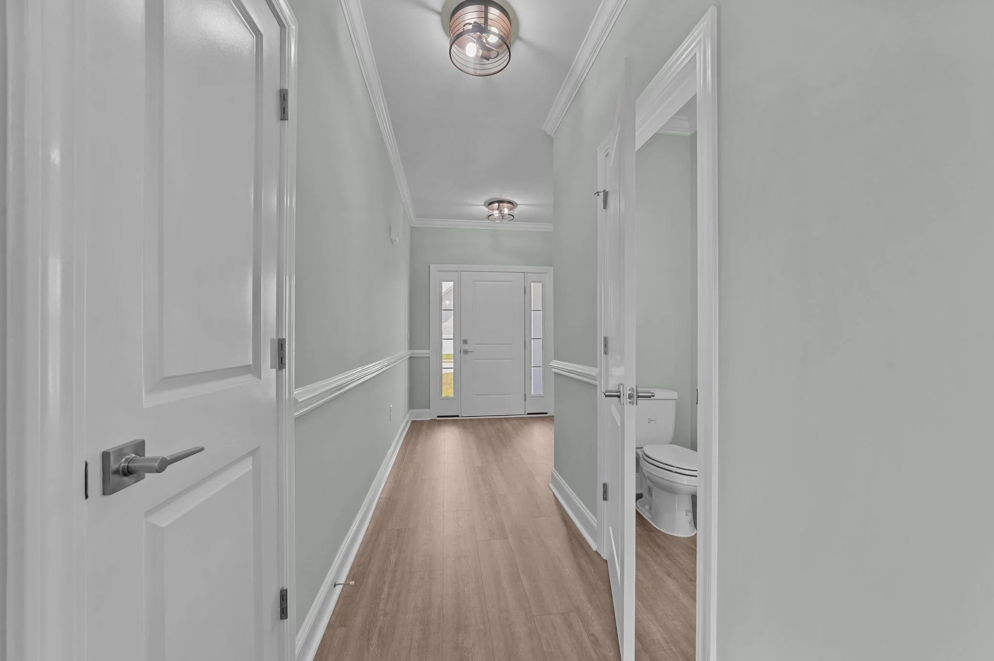 Interior view of a modern house hallway with white walls, wooden flooring, and ceiling lights; a partially open door reveals a small bathroom with a toilet.