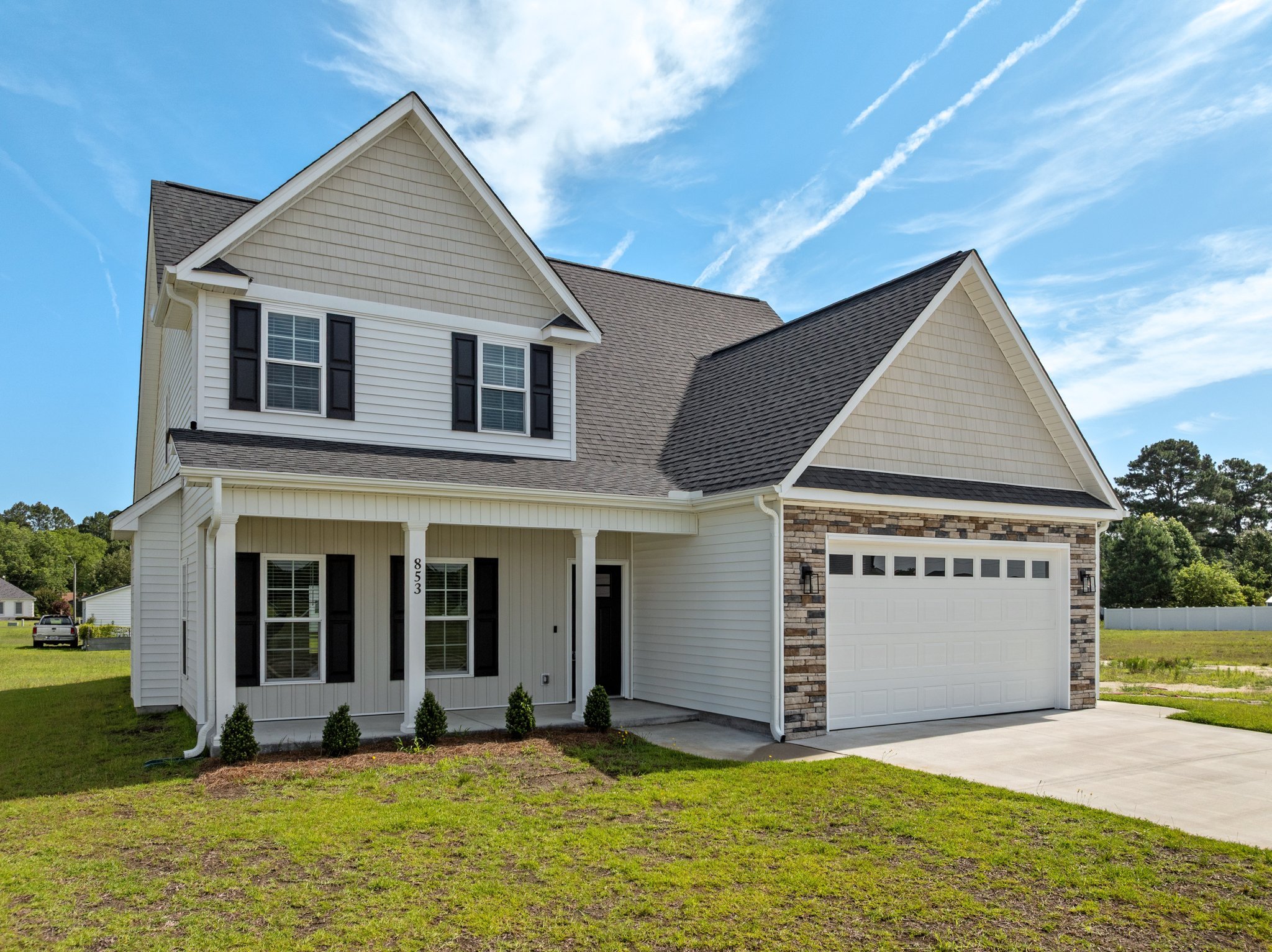 Front view of a modern two-story house with beige siding, black shutters, and a combination of brick and siding exterior, with a garage and a well-maintained lawn under a partly cloudy sky.