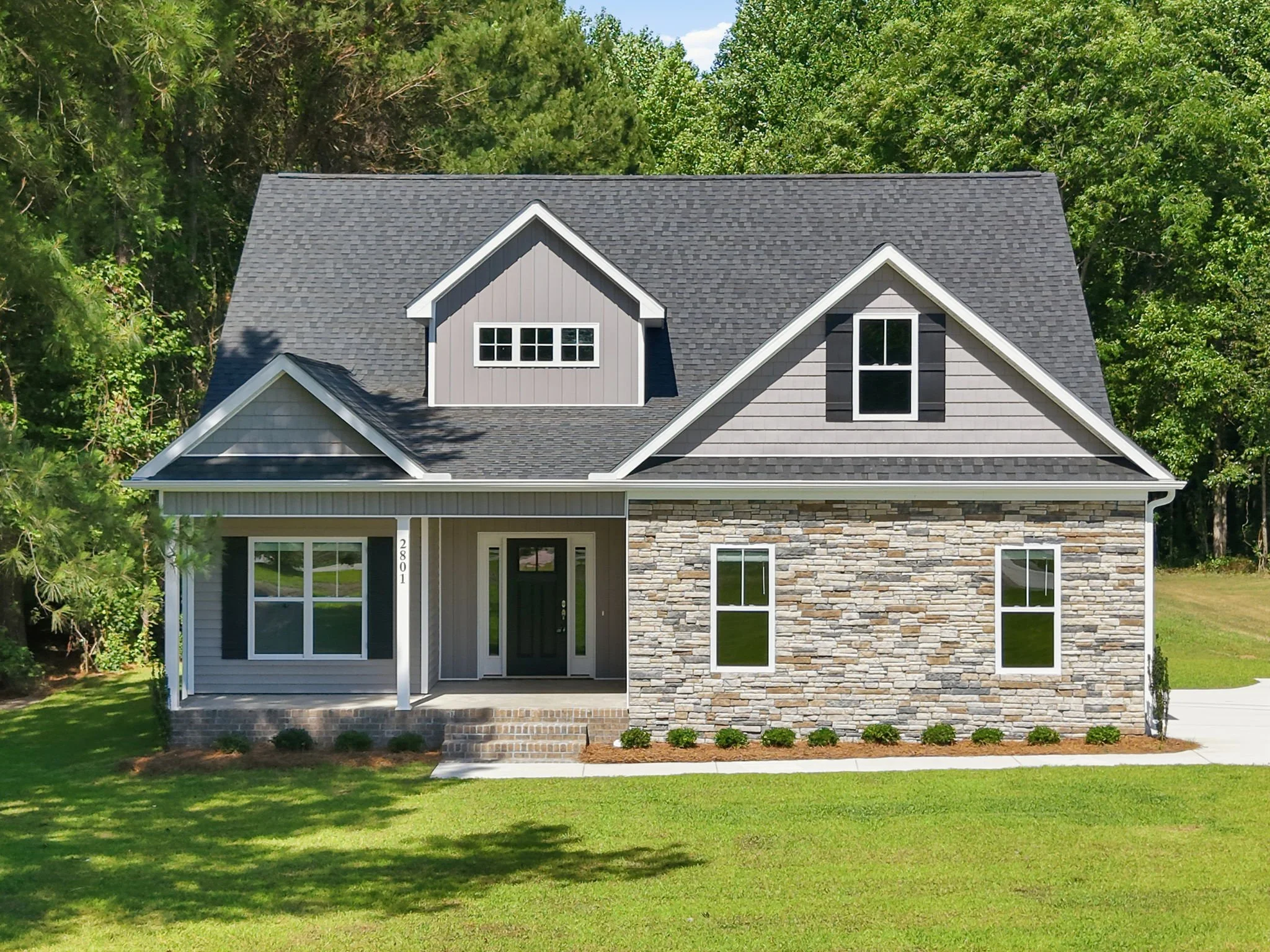 Modern two-story house with gray siding, stone accents, and multiple dormer windows, surrounded by a green lawn and trees.