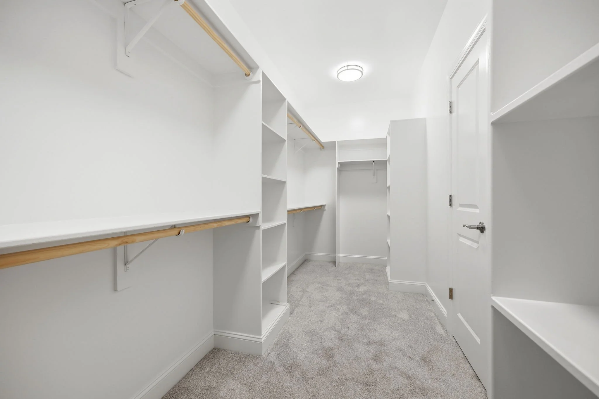 Empty walk-in closet with white walls and built-in shelves and rods for clothing, gray carpeted floor, and a ceiling light.