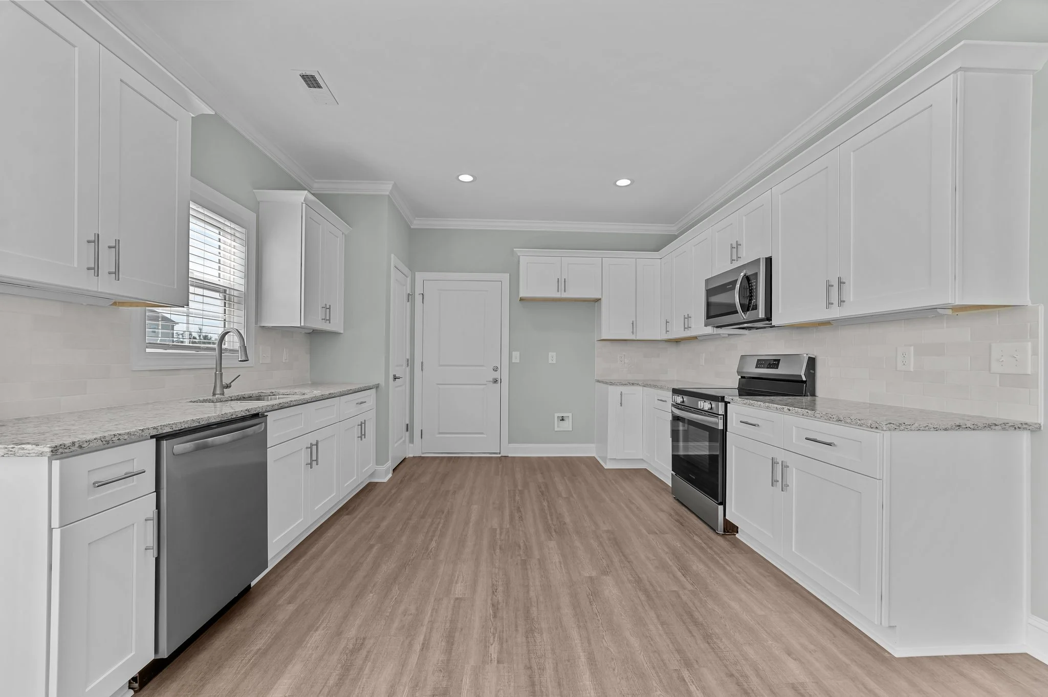 Modern kitchen with white cabinets, stainless steel appliances, granite countertops, wood flooring, and a window with blinds.