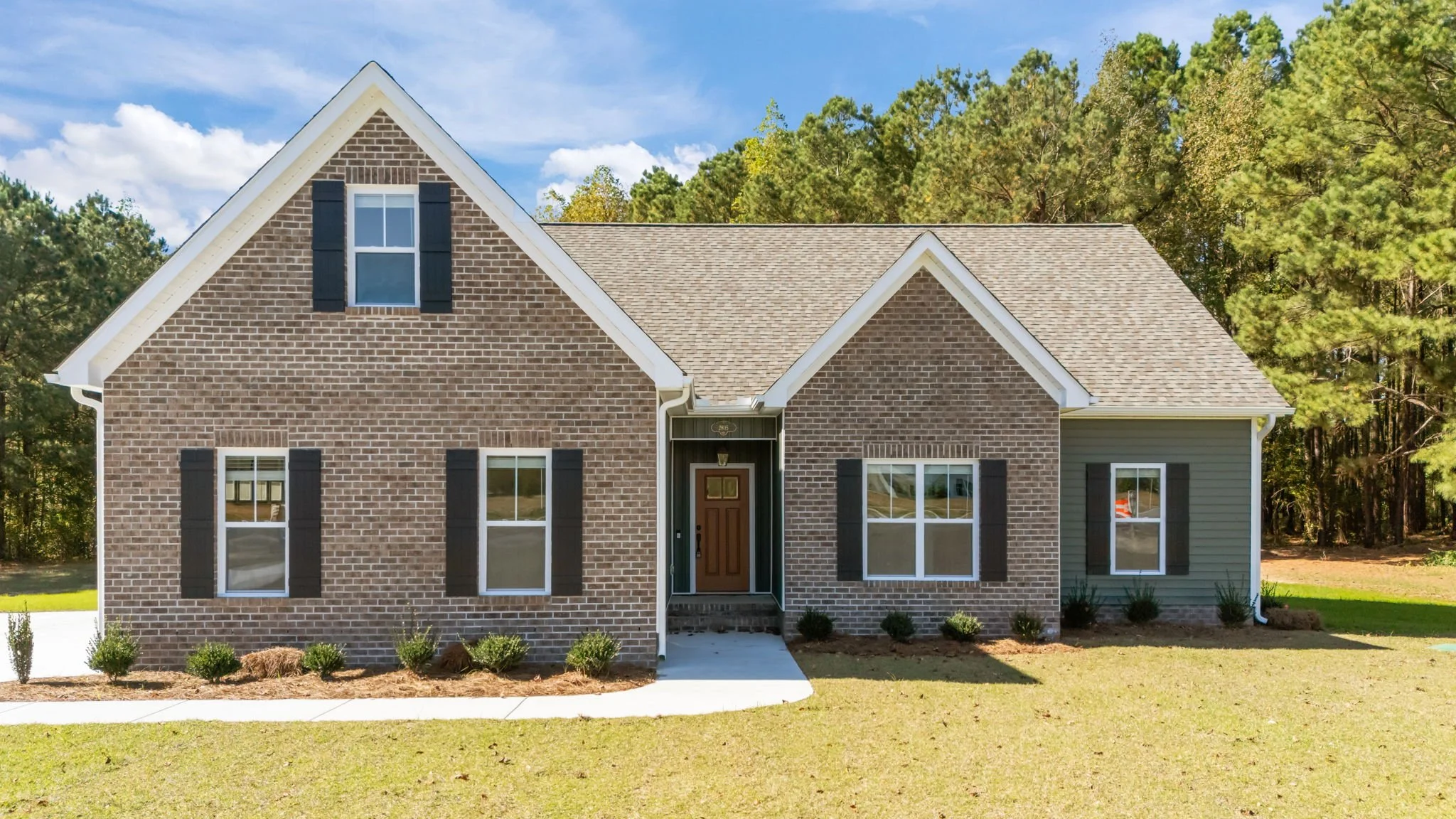 Front view of a two-story house with brick and gray siding, multiple windows with black shutters, and a brown front door, surrounded by a green lawn and trees under a partly cloudy sky.