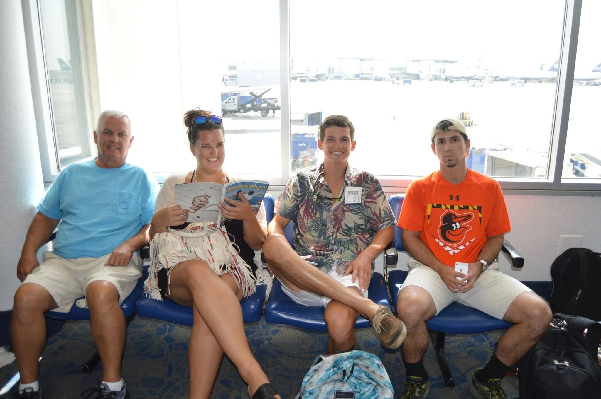 Four people sitting in an airport waiting area with large windows in the background showing airplanes and airport tarmac. The group includes an older man in a blue shirt, a woman with sunglasses on her head reading a brochure, a young man in a Hawaiian shirt, and a young man in an orange T-shirt wearing a cap. They are seated in blue chairs with bags on the floor nearby.