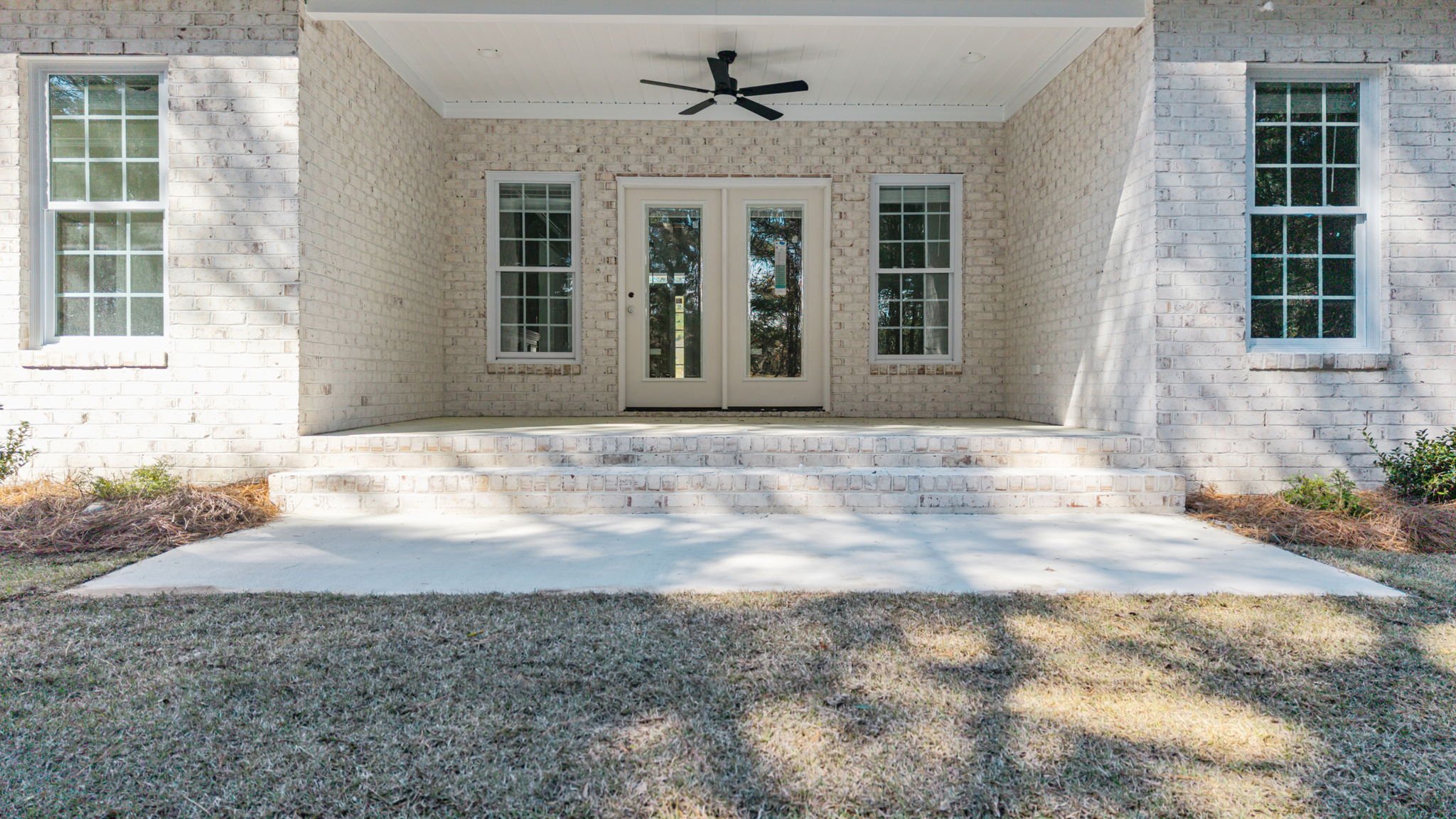 Back porch with brick walls, white double doors, two windows, concrete steps, and a ceiling fan.