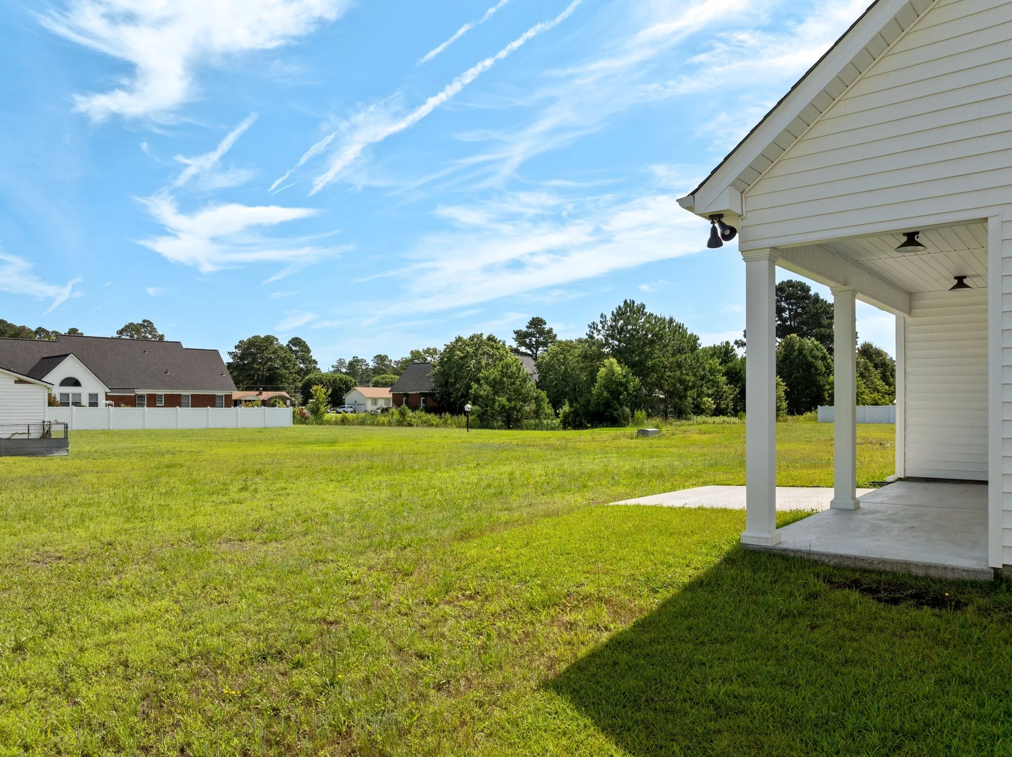 View of a backyard with a white house porch, green grassy yard, and neighboring houses with trees under a bright blue sky with wispy clouds.