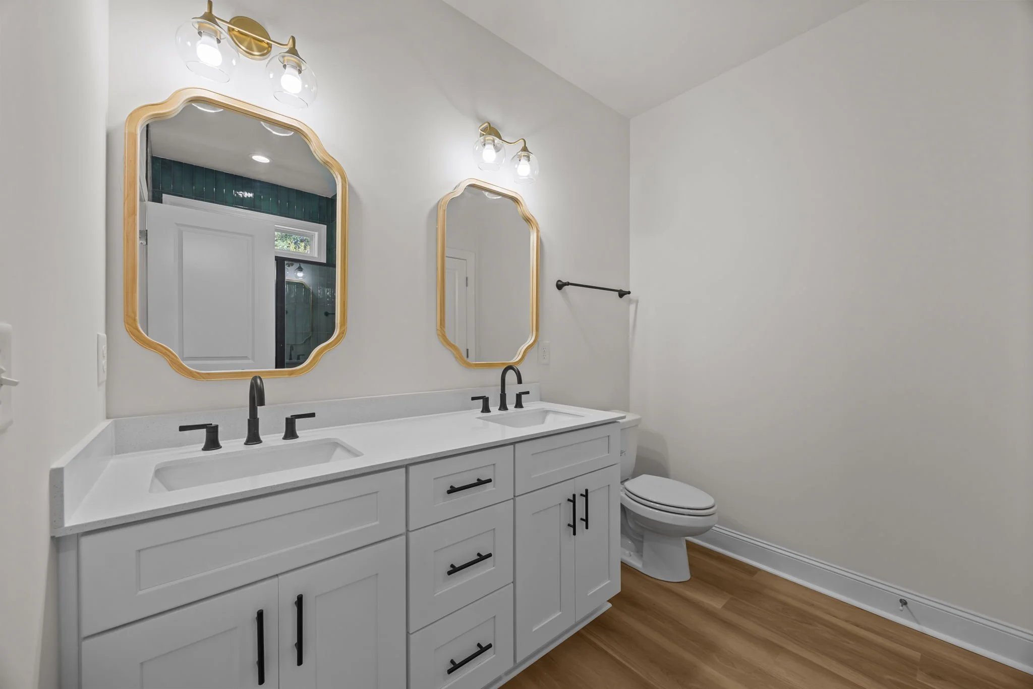 A modern bathroom with two white sinks, black faucets, and a white vanity with black handles. Above the sinks are two ornate framed mirrors, each illuminated by a light fixture with three bulbs. To the right of the vanity is a white toilet, and the f