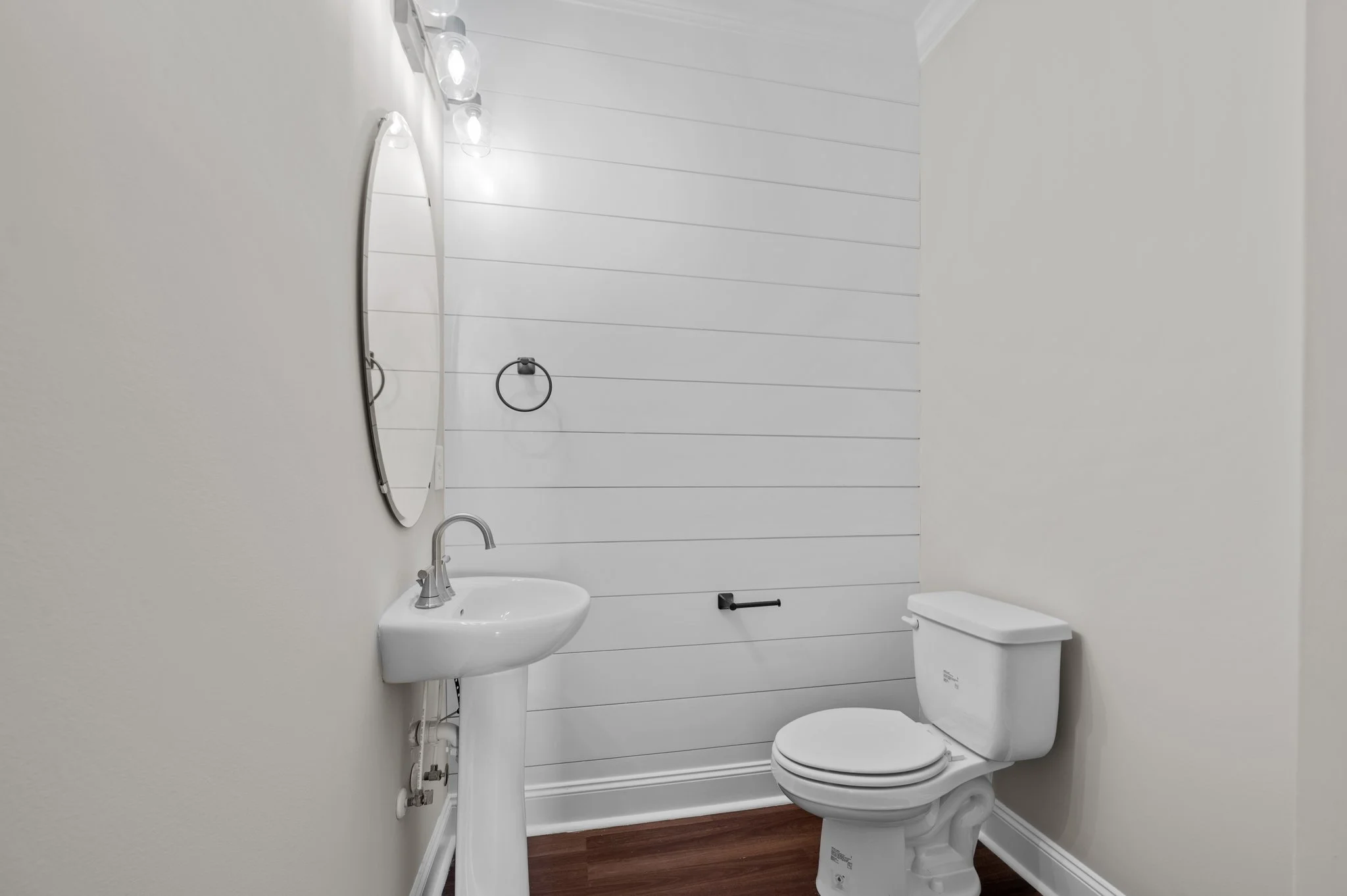 A small bathroom with white walls and shiplap paneling on one wall. It contains a white toilet, a pedestal sink, and a mirror. There are three wall-mounted lights above the mirror and a towel ring next to it. The floor is wood.
