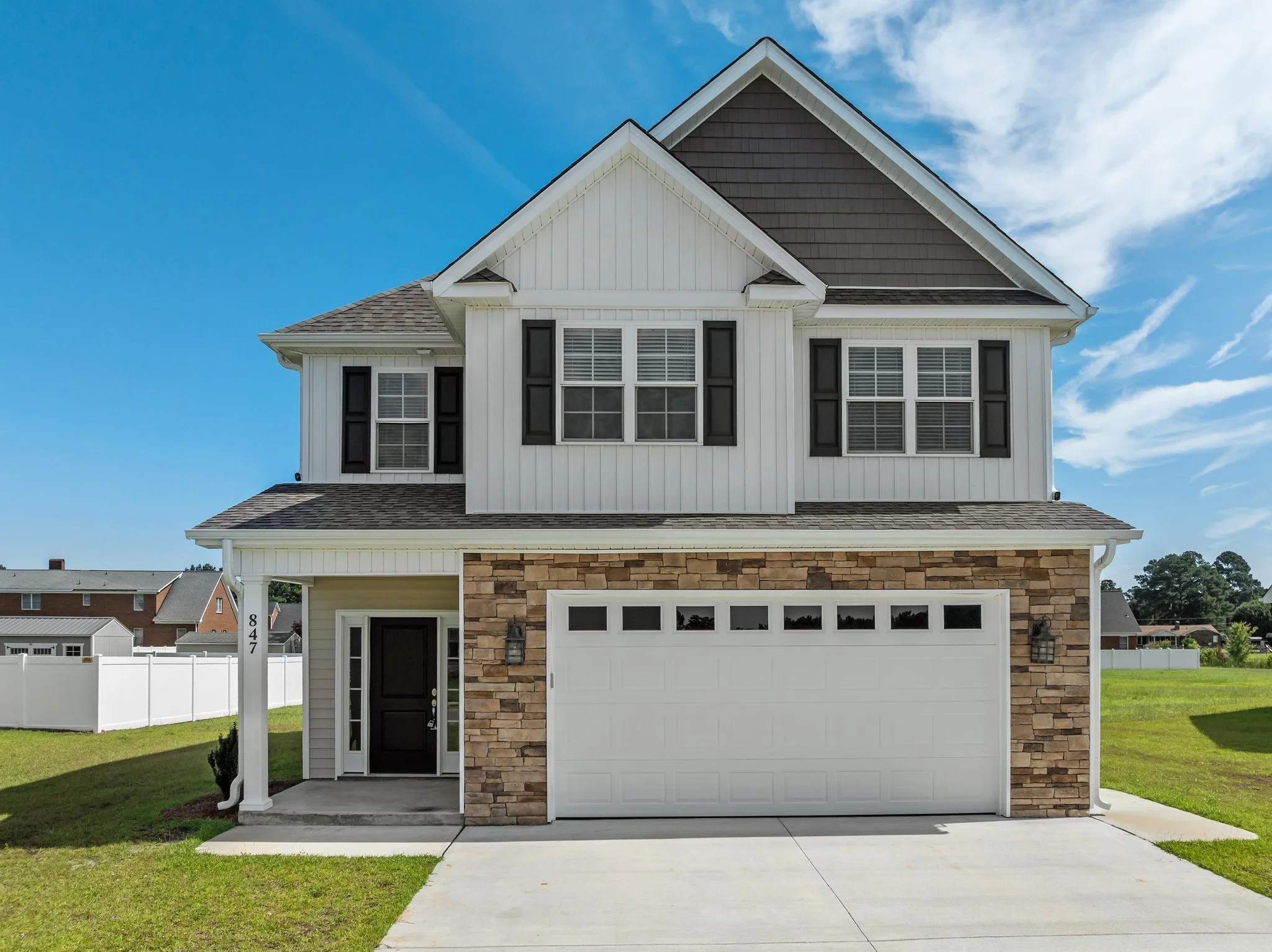 Front view of a modern two-story house with brick and white siding, a white garage door, black shutters, and a black front door. The house has a gabled roof and is surrounded by a green lawn. White fence is visible in the background under a partly cloudy sky.