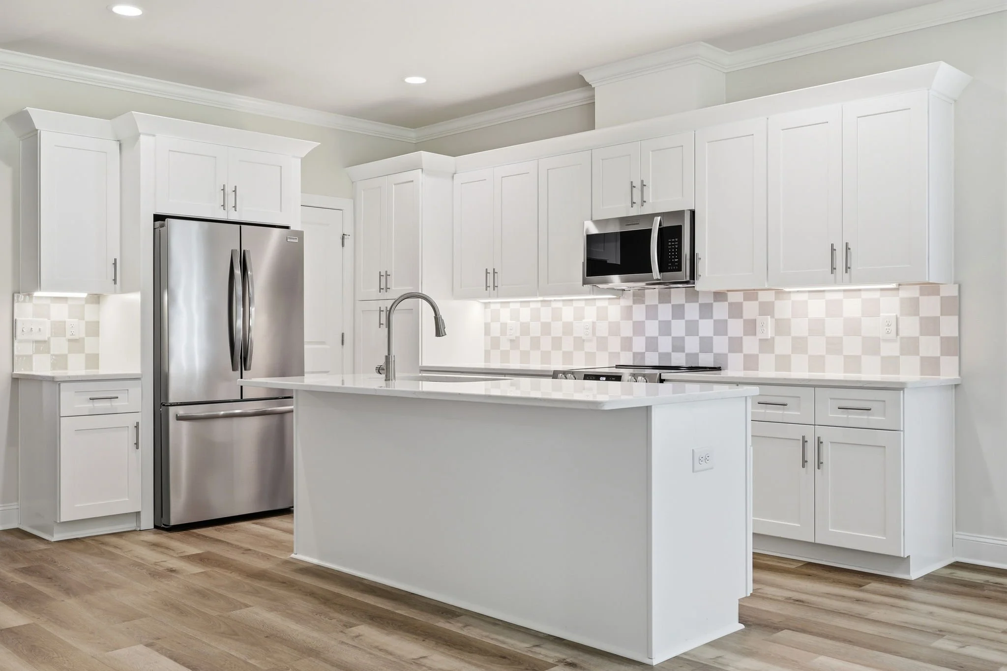 Modern white kitchen with stainless steel refrigerator, microwave, and island with sink. Wooden flooring and checkered backsplash.