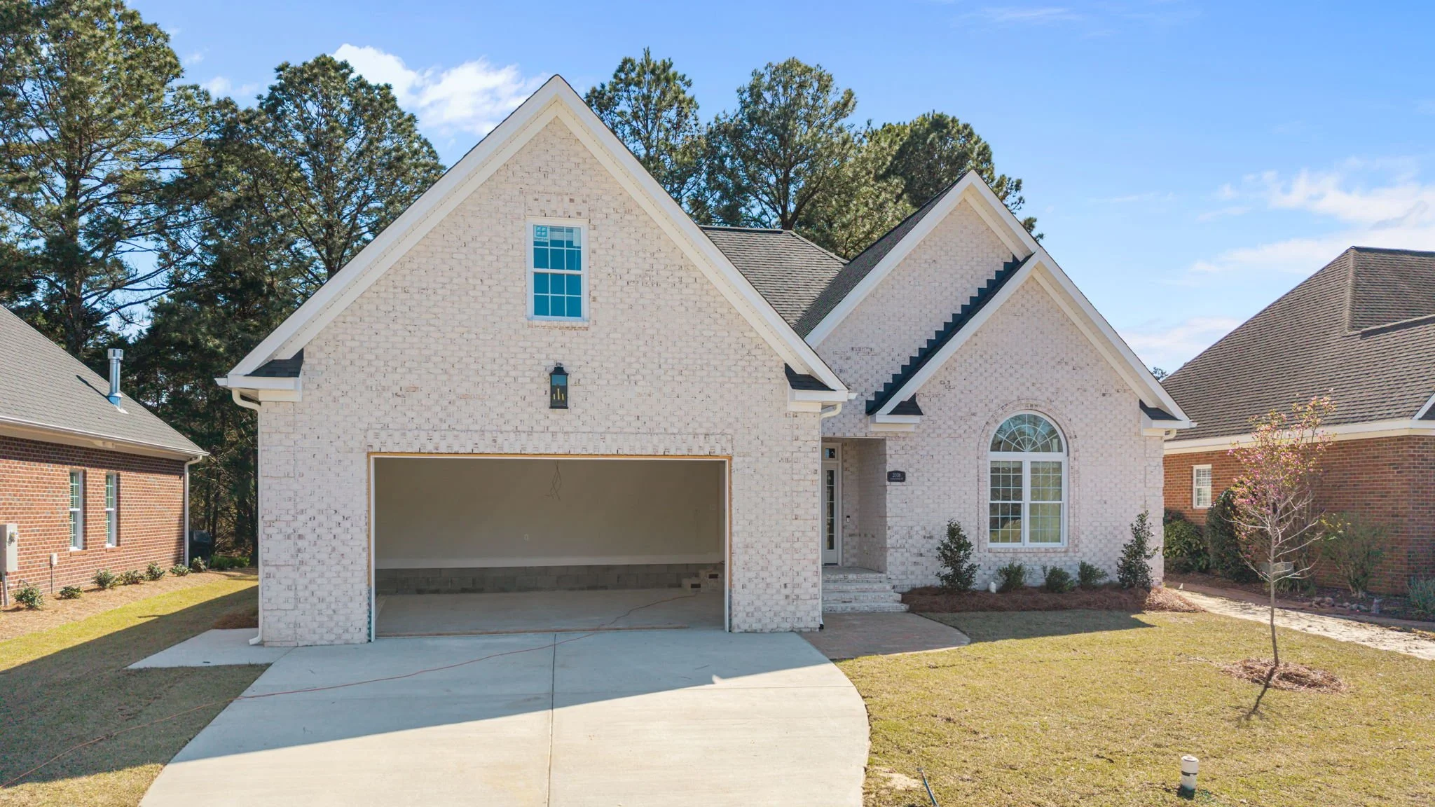 New two-story house with white brick exterior, large arched window, and an attached garage under construction, on a lawn with a small tree under a blue sky.