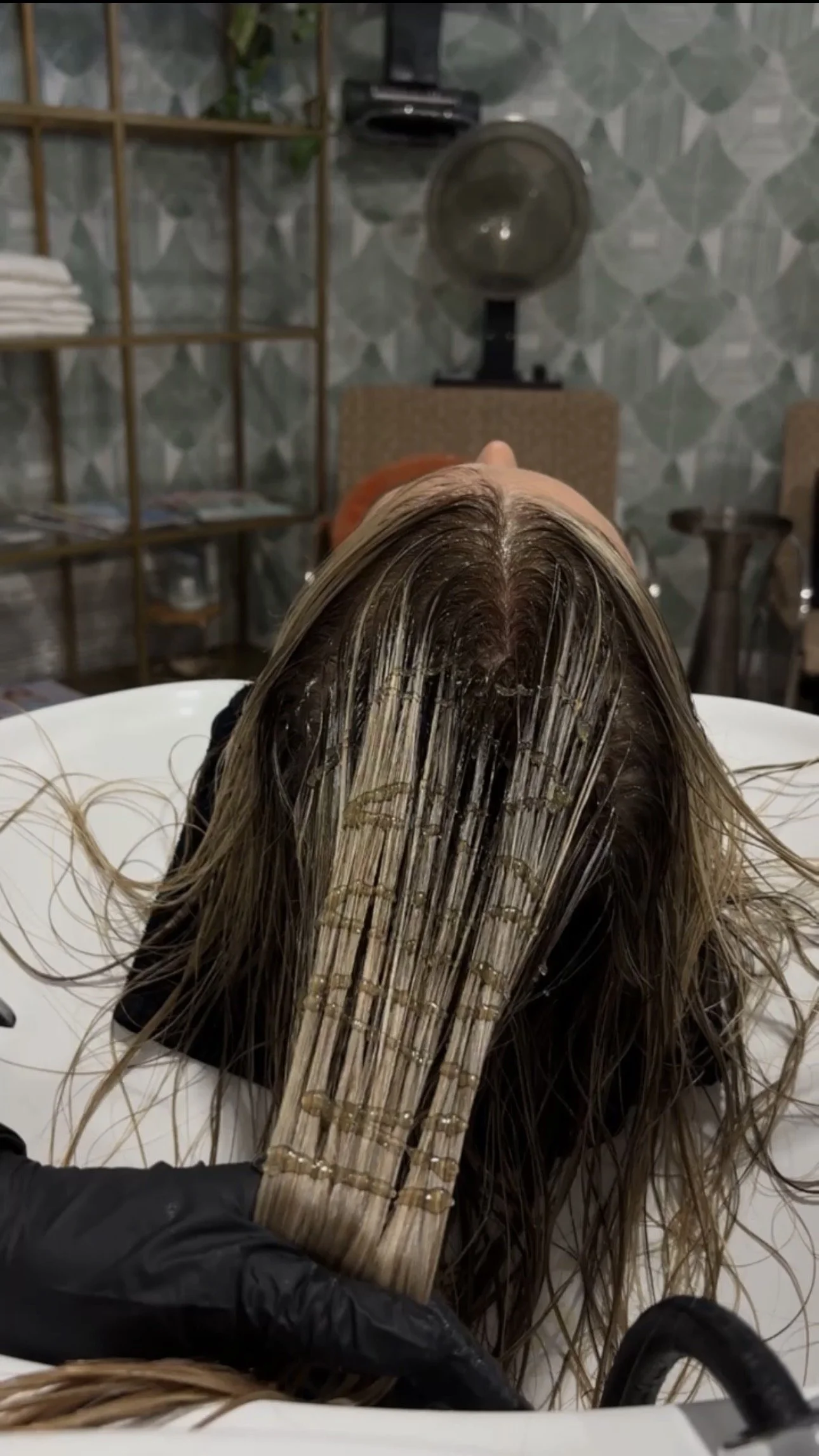A person is reclining with their hair being treated, with hair strands separated and oil applied, in a salon setting, with a hairdryer and salon furniture visible in the background.